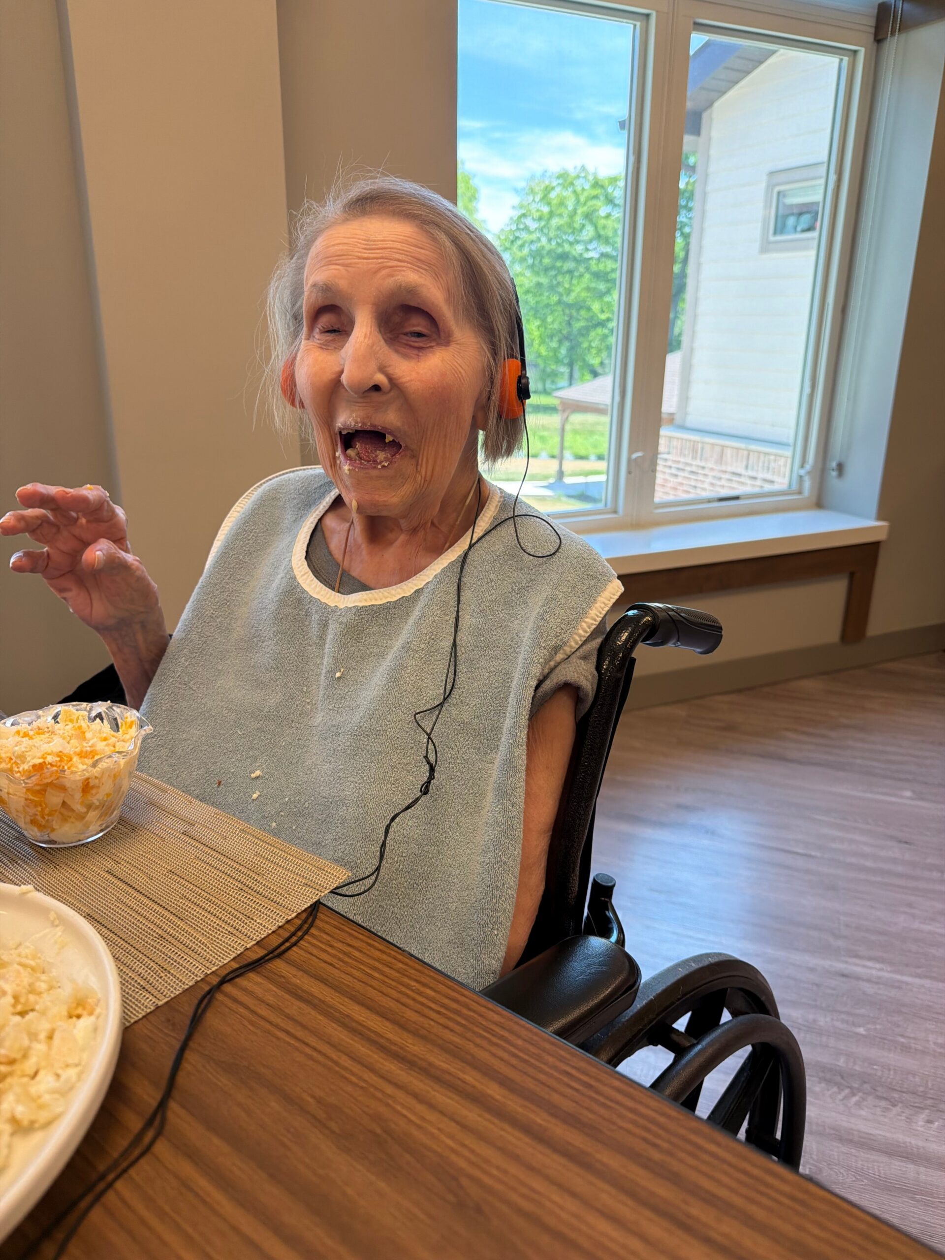 Older woman in a wheelchair wearing a light blue bib and orange headphones, smiling while eating macaroni salad at a dining table by a window.