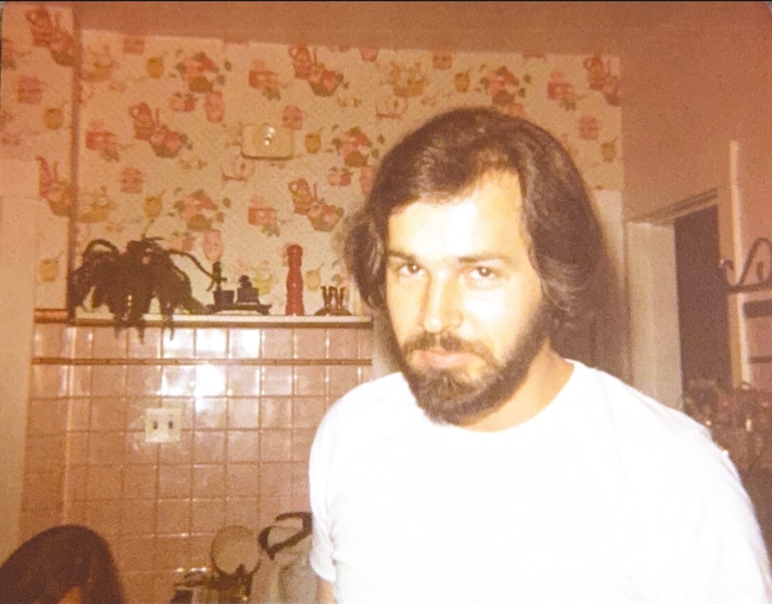 Bearded man in a white shirt standing in a retro kitchen with pink tile backsplash and floral wallpaper.