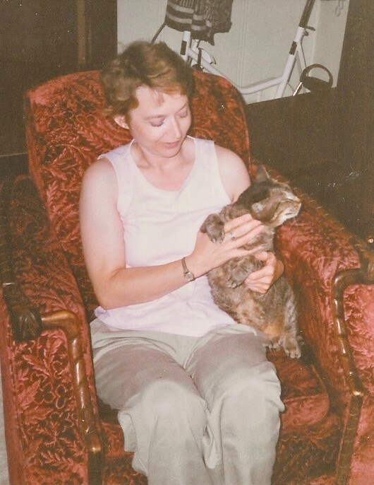 Woman in a white tank top seated in a red patterned armchair, gently holding a small dog.