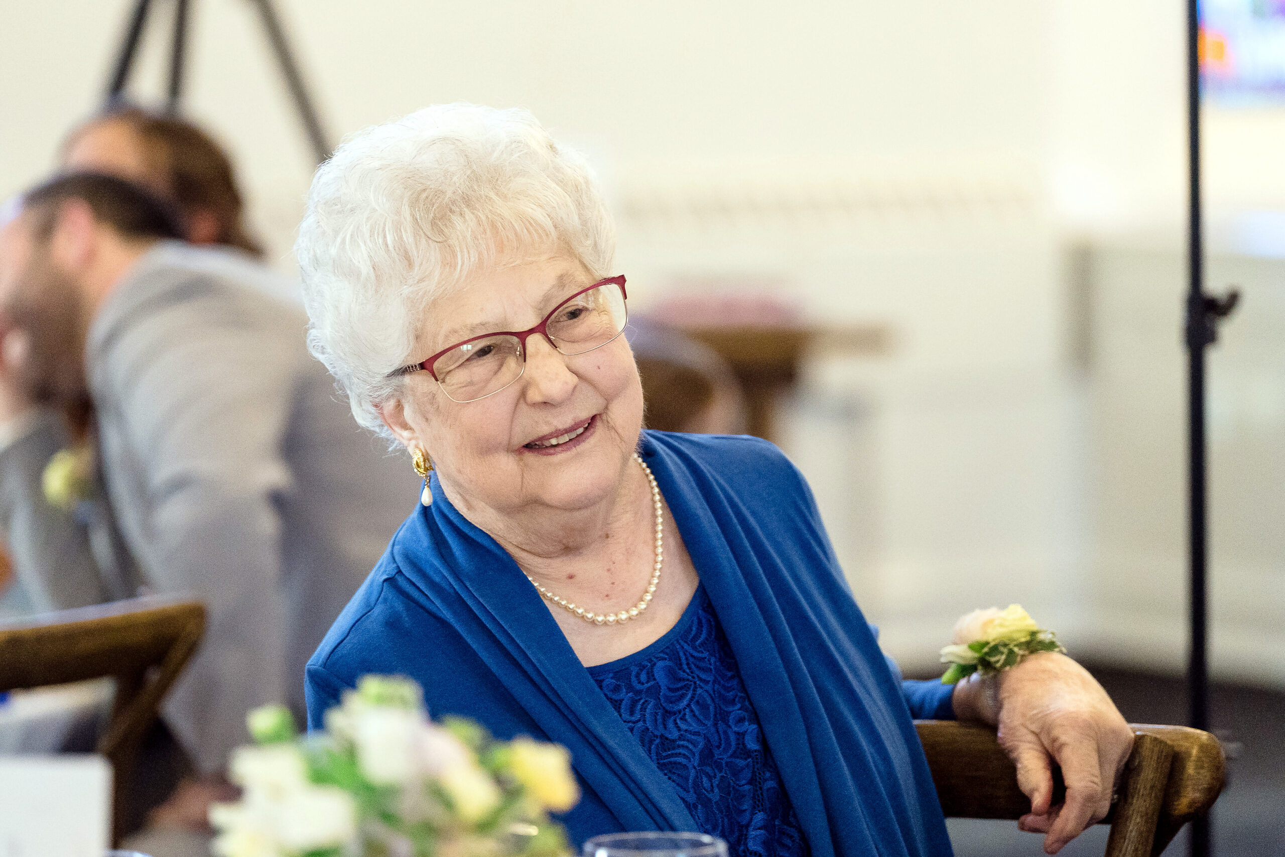 Smiling elderly woman with short white hair and red glasses, wearing blue attire at a social gathering.