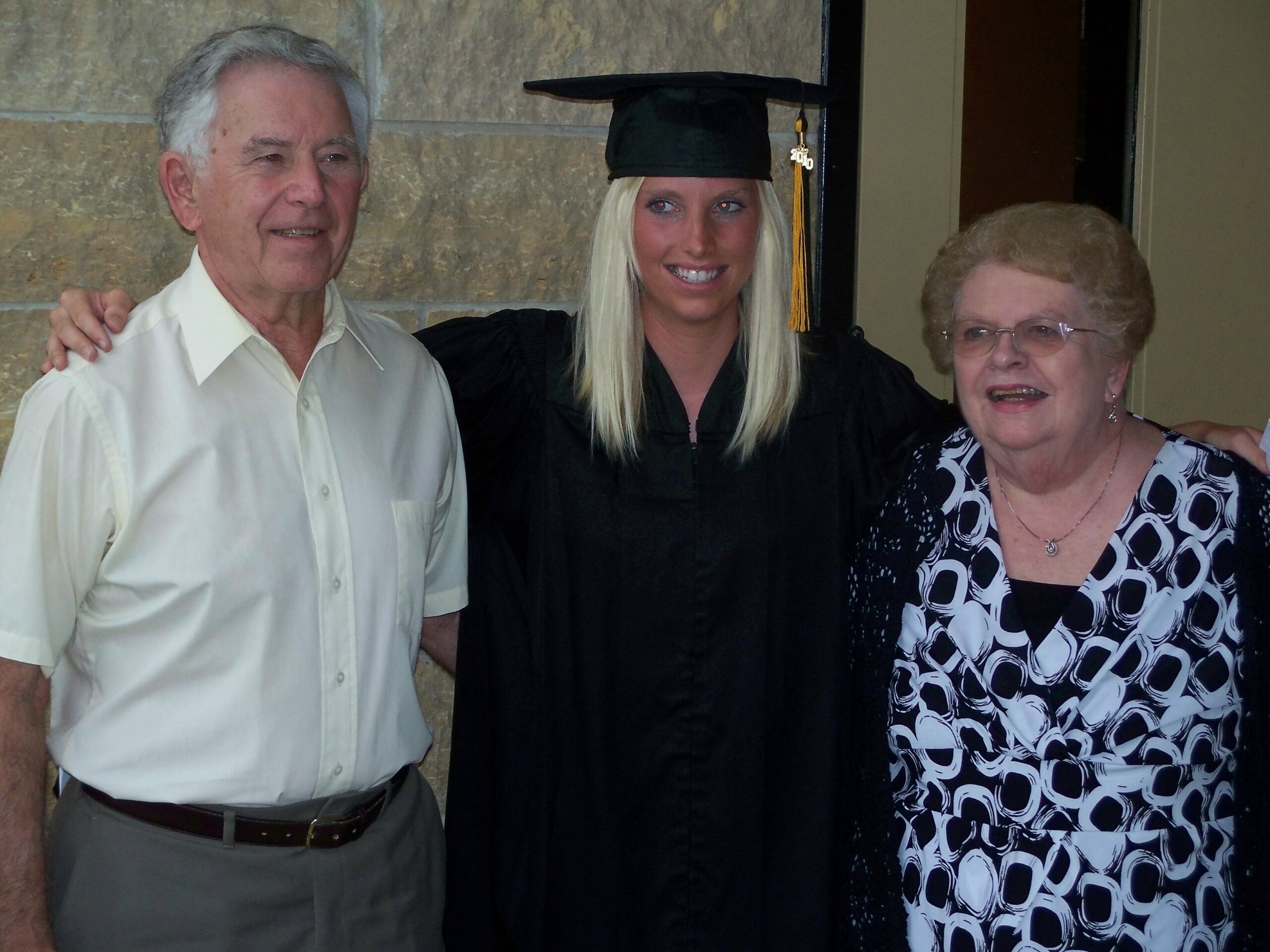 Three people posing indoors for a graduation photo: a graduate in cap and gown flanked by an older man in a white shirt and an older woman in a patterned blouse, all smiling.