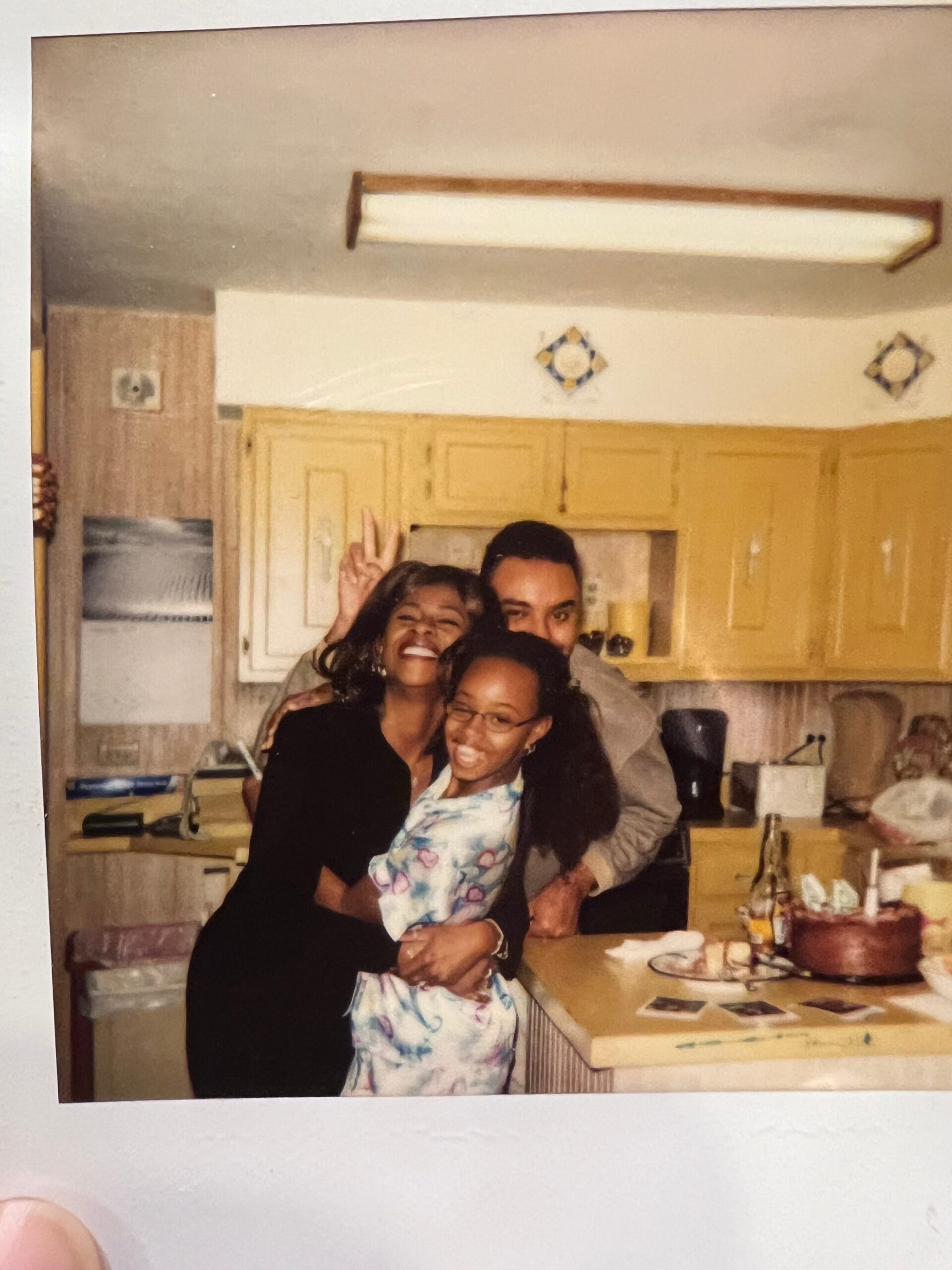 Four friends posing and smiling in a kitchen, one flashing a peace sign on the left side of the photo.