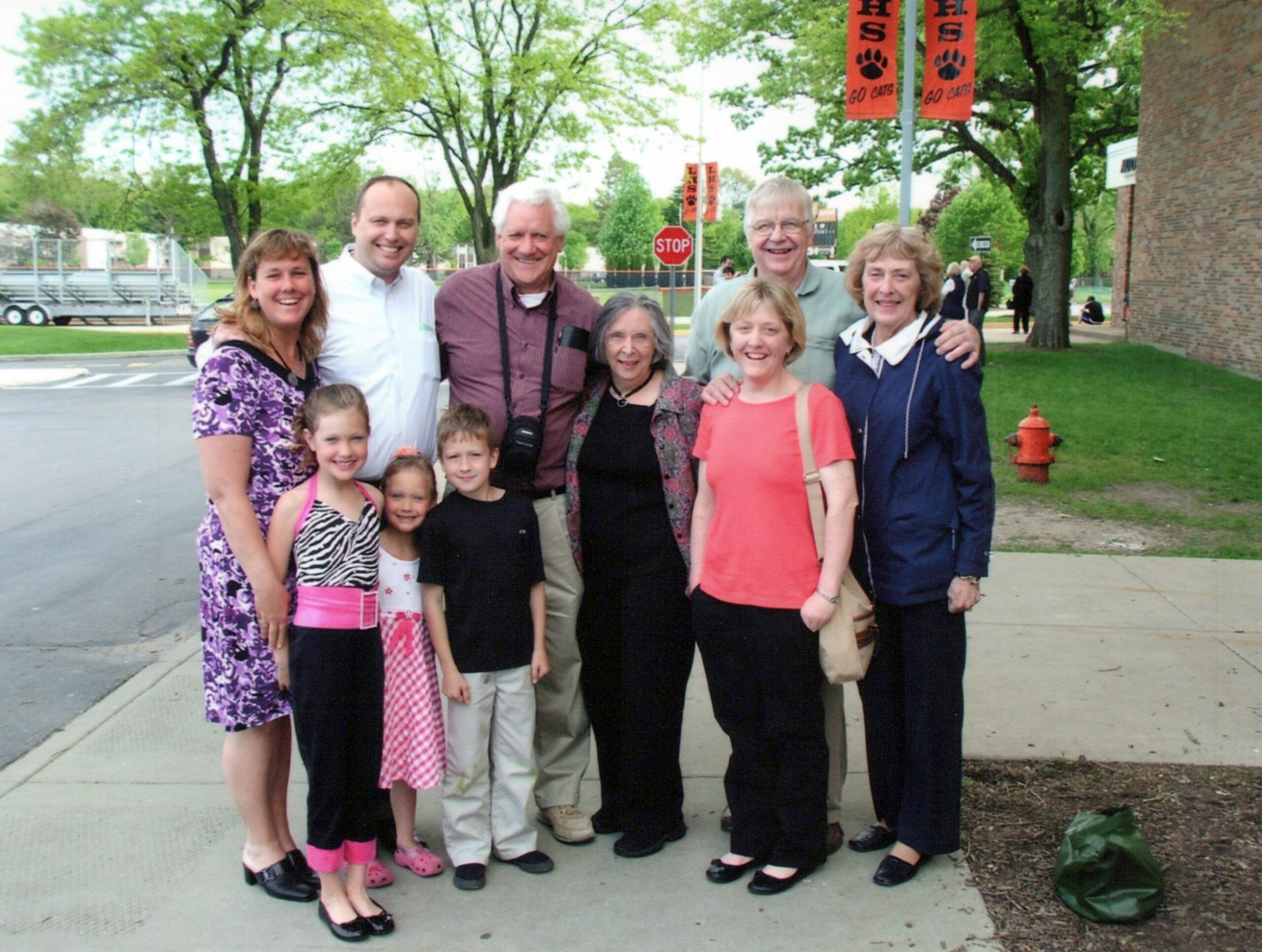 A group of smiling adults and children posing together outdoors on a sidewalk with trees and orange banners in the background.