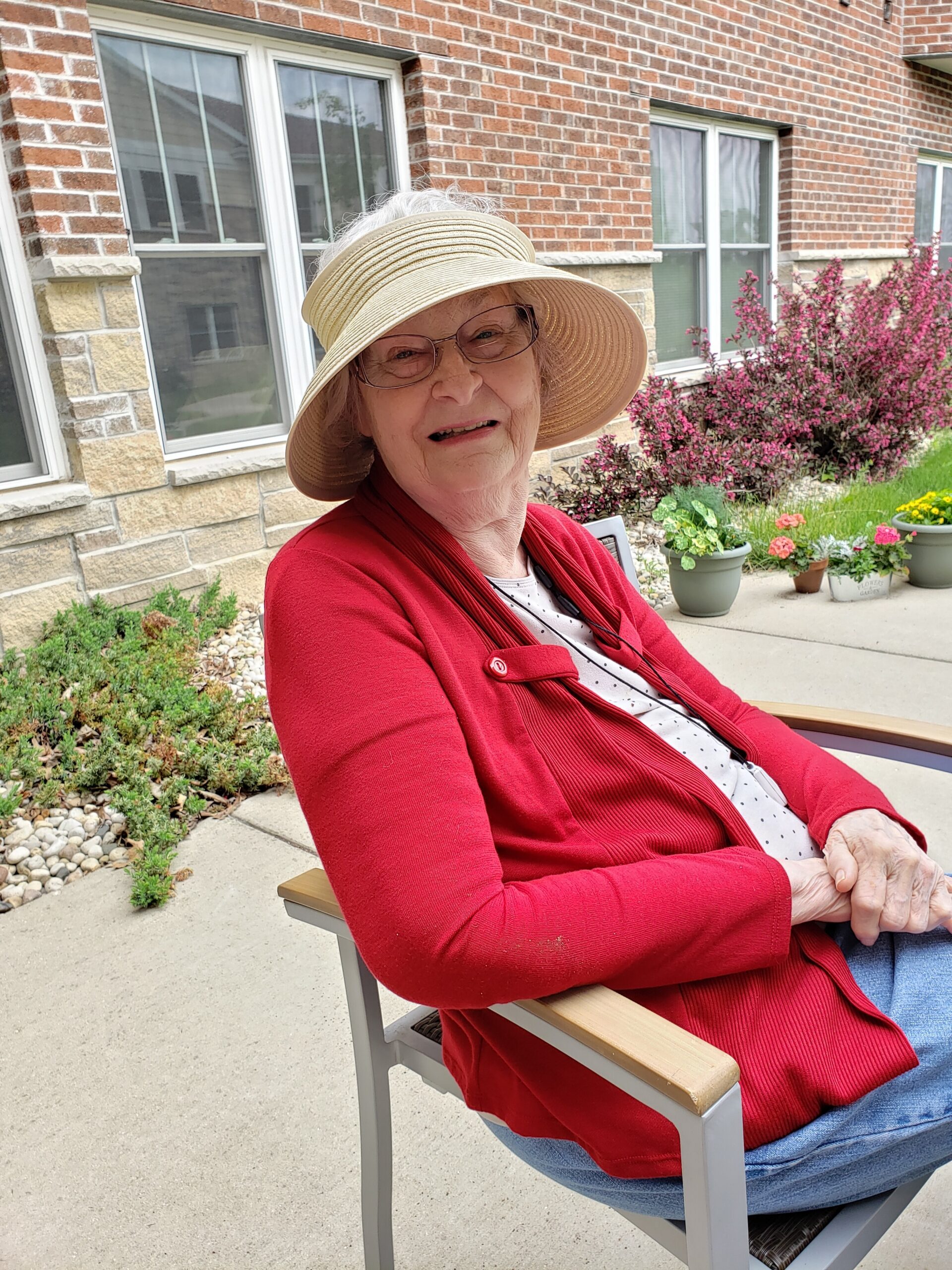 An elderly woman wearing a wide-brim hat and red cardigan sits on a chair outside a brick building with flowering bushes in the background.