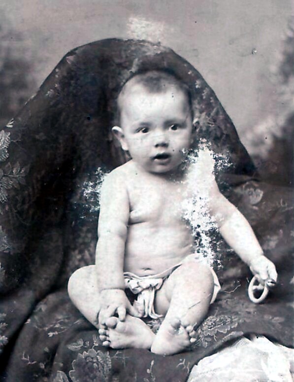 Black-and-white portrait of a baby sitting up on patterned fabric, wearing a diaper and holding a teething ring in one hand, facing the camera.