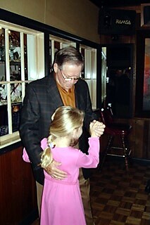 Older man in a blazer dancing with a young girl in a pink dress, holding hands in a dim bar or restaurant setting.