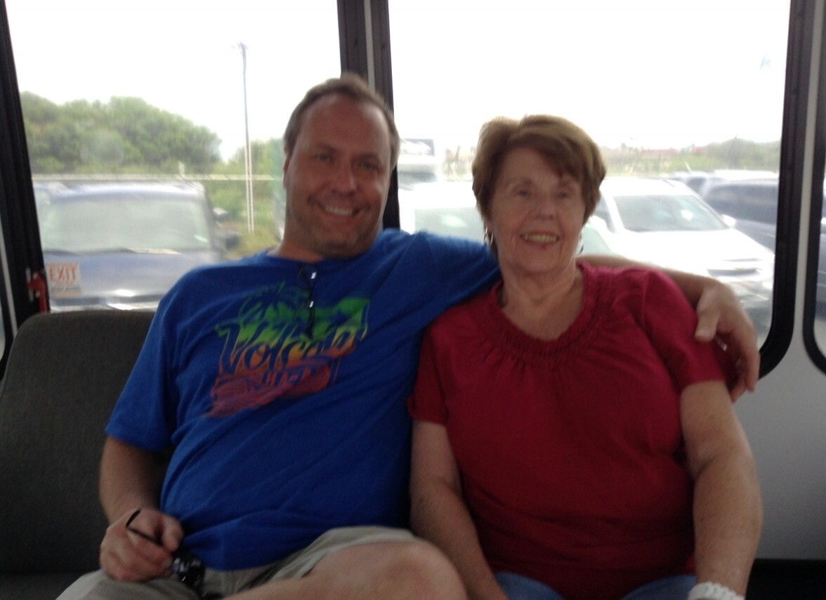 Man and an older woman sit close together on a bus, smiling at the camera; the man wears a blue T-shirt with a palm tree graphic, the woman wears a red blouse, and their arms are around each other. Outside the windows are parked cars and greenery.
