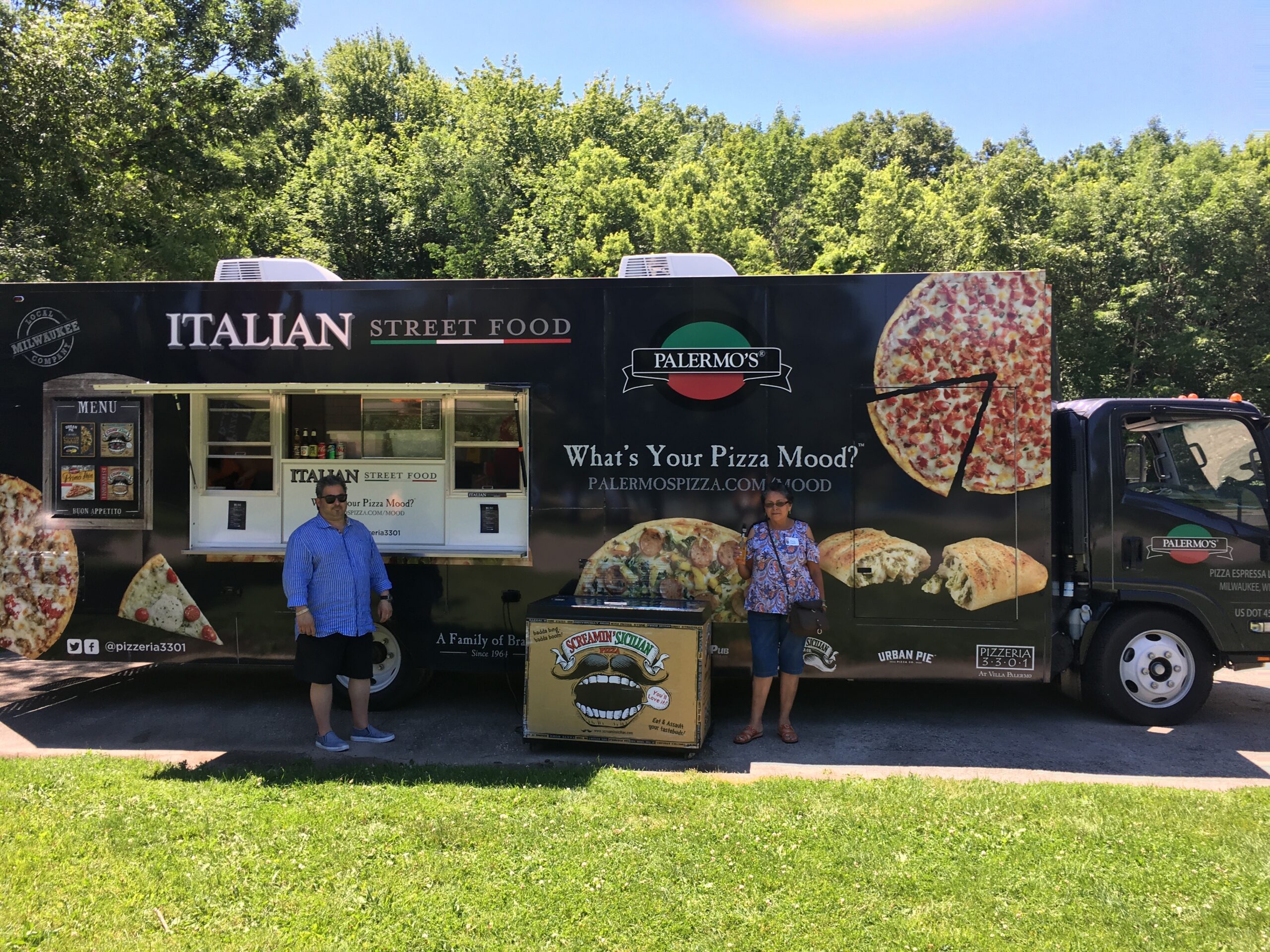 Pizza truck labeled Italian Street Food with Palermo's branding and large pizza images on the side; two people stand in front, grassy park setting behind trees.