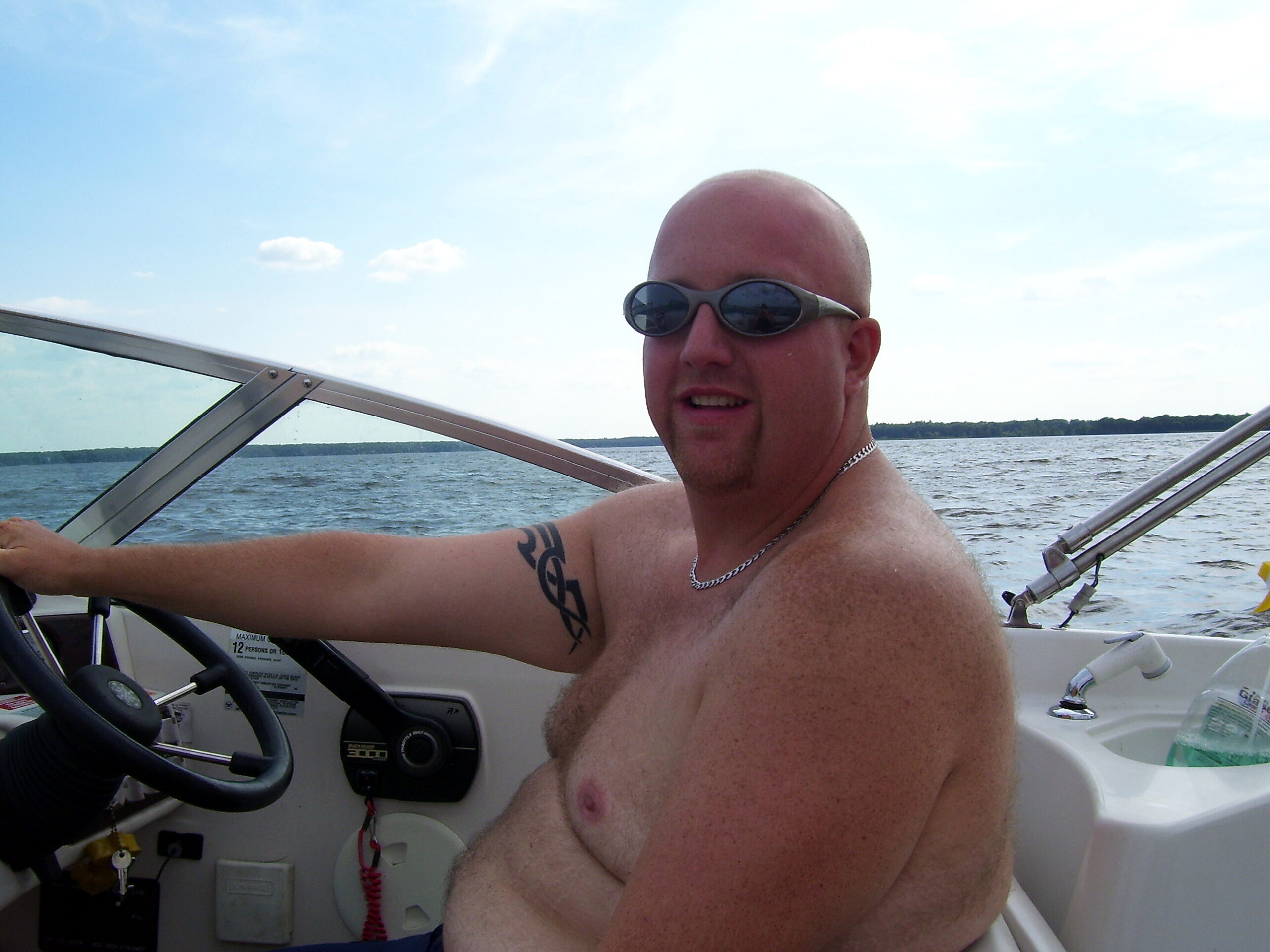 Man with a shaved head and sunglasses sits at the helm of a boat on a lake under a blue sky.