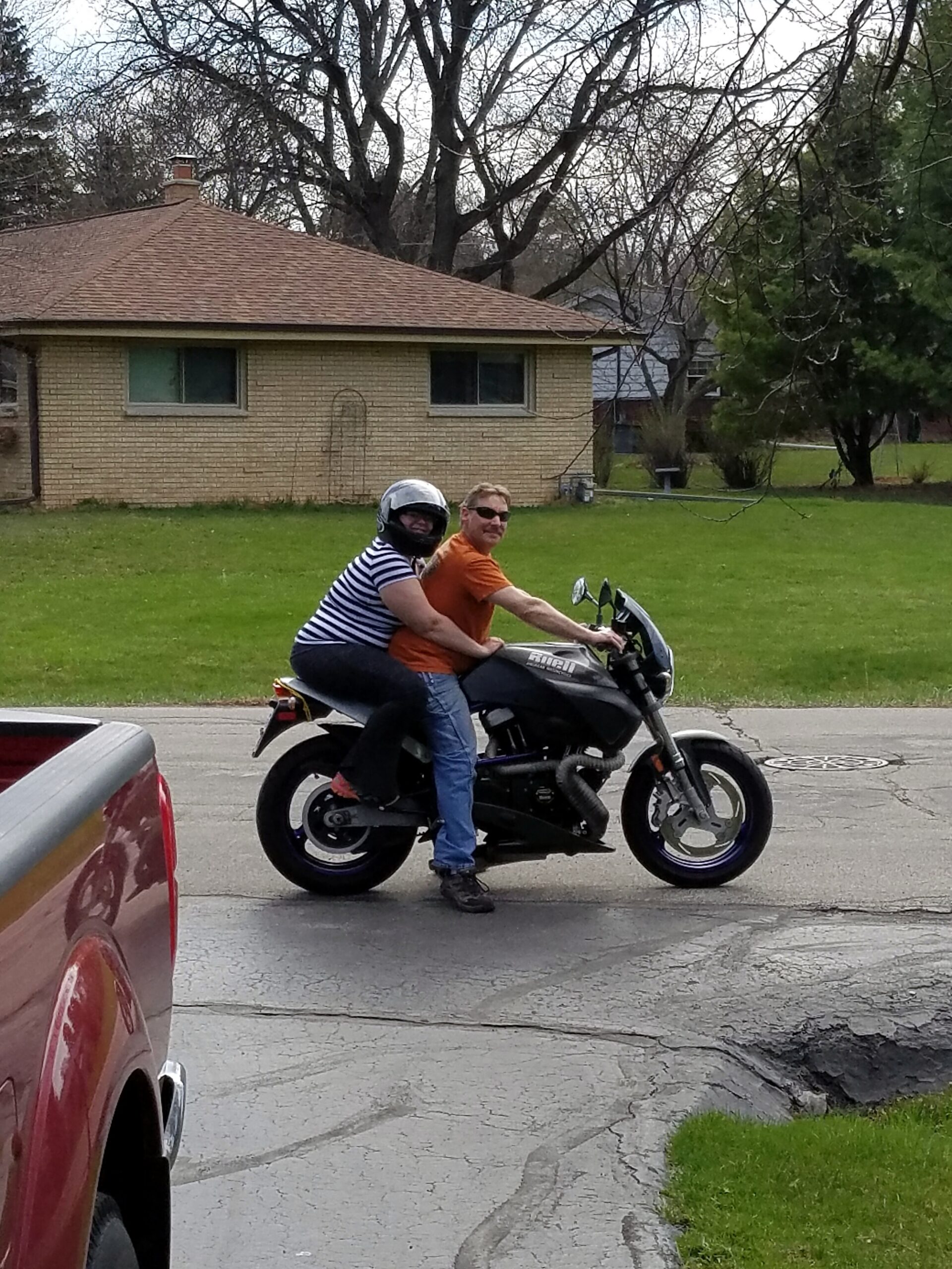 Two people ride a motorcycle on a suburban street, with a brick house and leafless trees in the background.