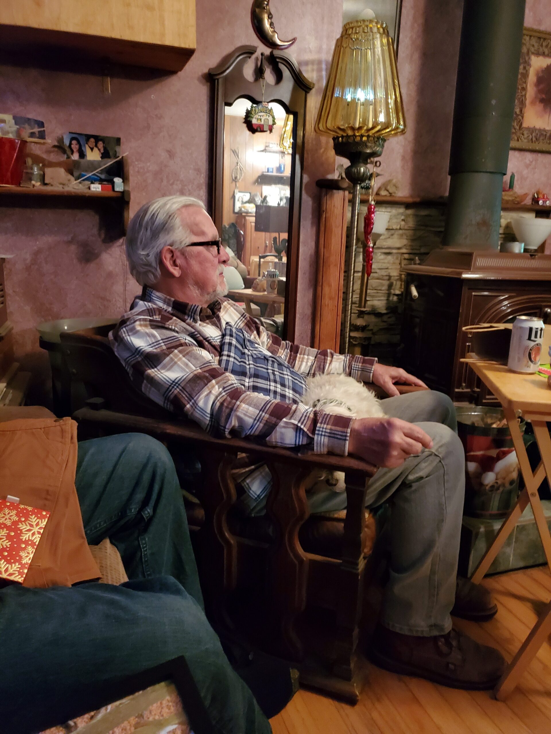 Older man with gray hair and glasses sits in a wooden chair in a cozy living room, wearing a plaid shirt and watching something to his left.