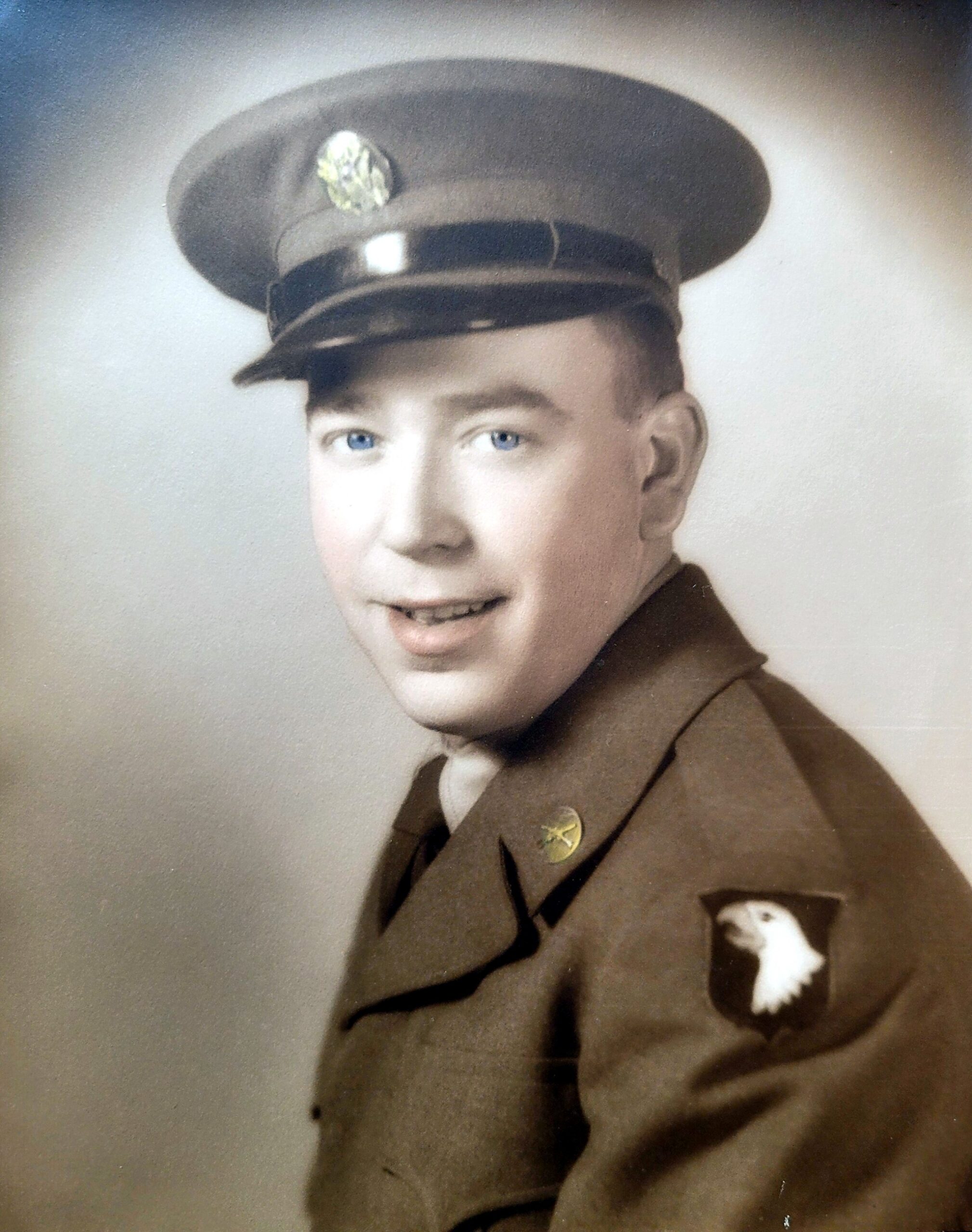 Young man in a vintage military uniform with a peaked cap, smiling; eagle insignia on the pocket