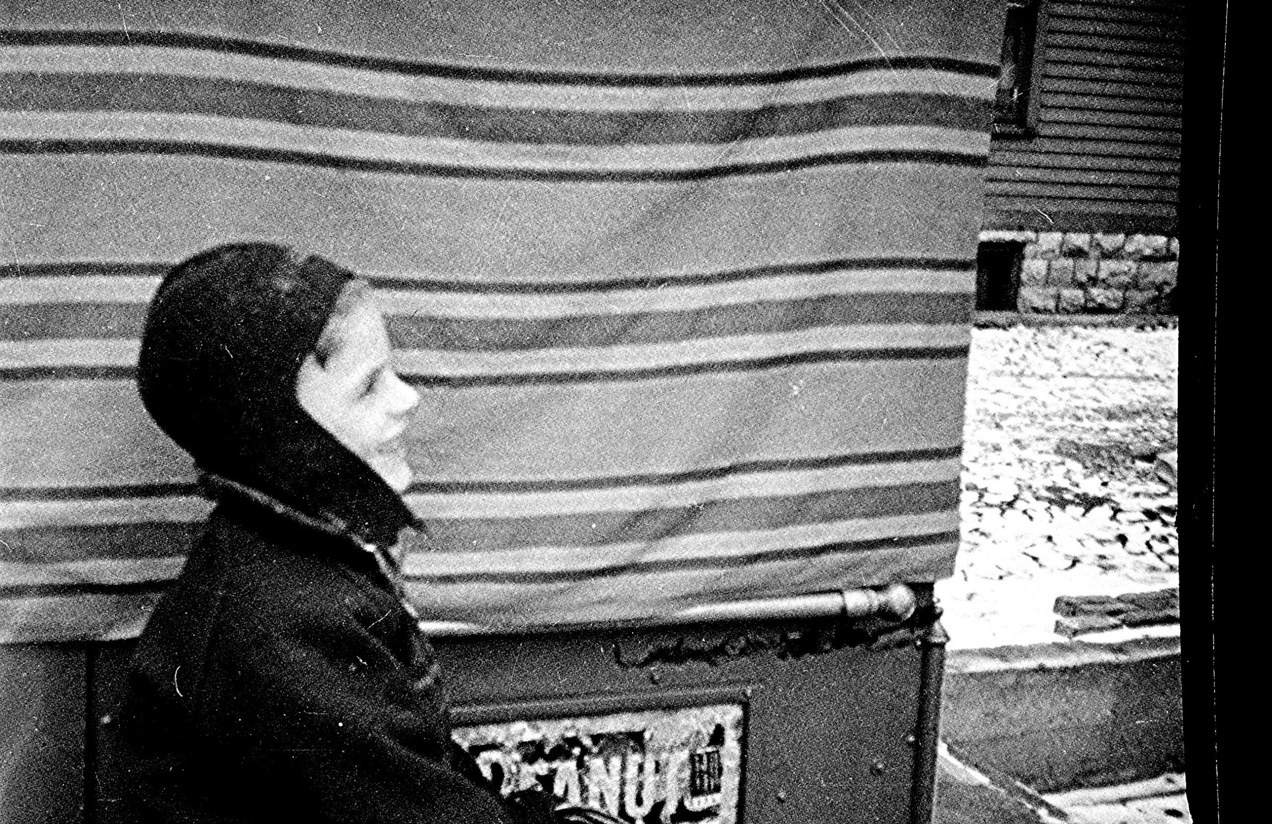 Black-and-white photo of a person in a hooded jacket standing beside a striped wall and a brick pillar, looking downward to the left.