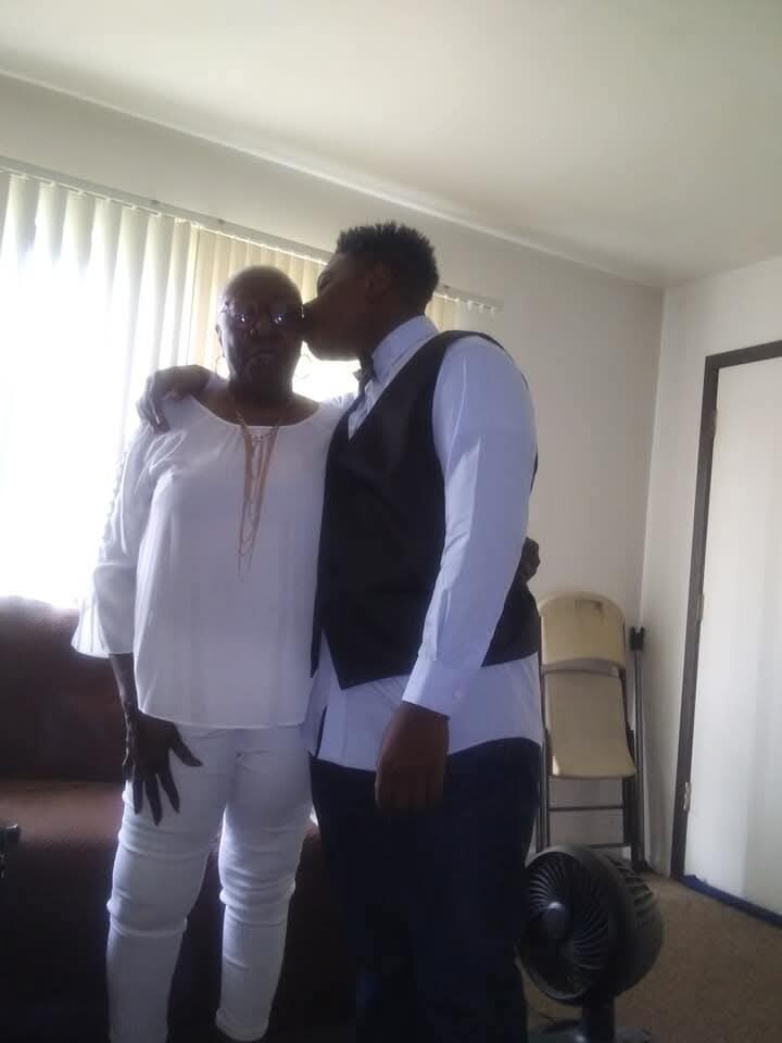 Two men stand close in a living room; one in white traditional clothing with sunglasses, the other in a white shirt and vest kissing his temple on the cheek.