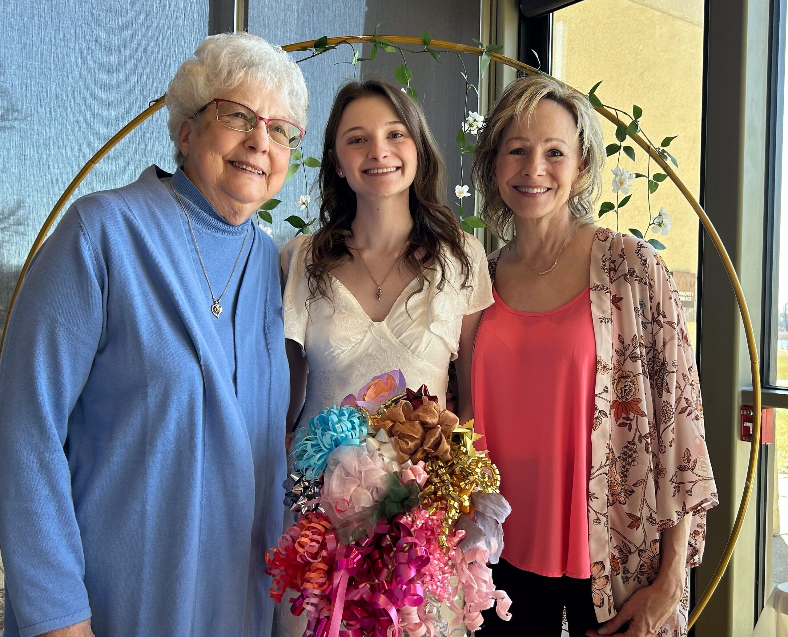 Three smiling women posing together indoors, standing in front of a circular plant arch with leaves and white flowers.
