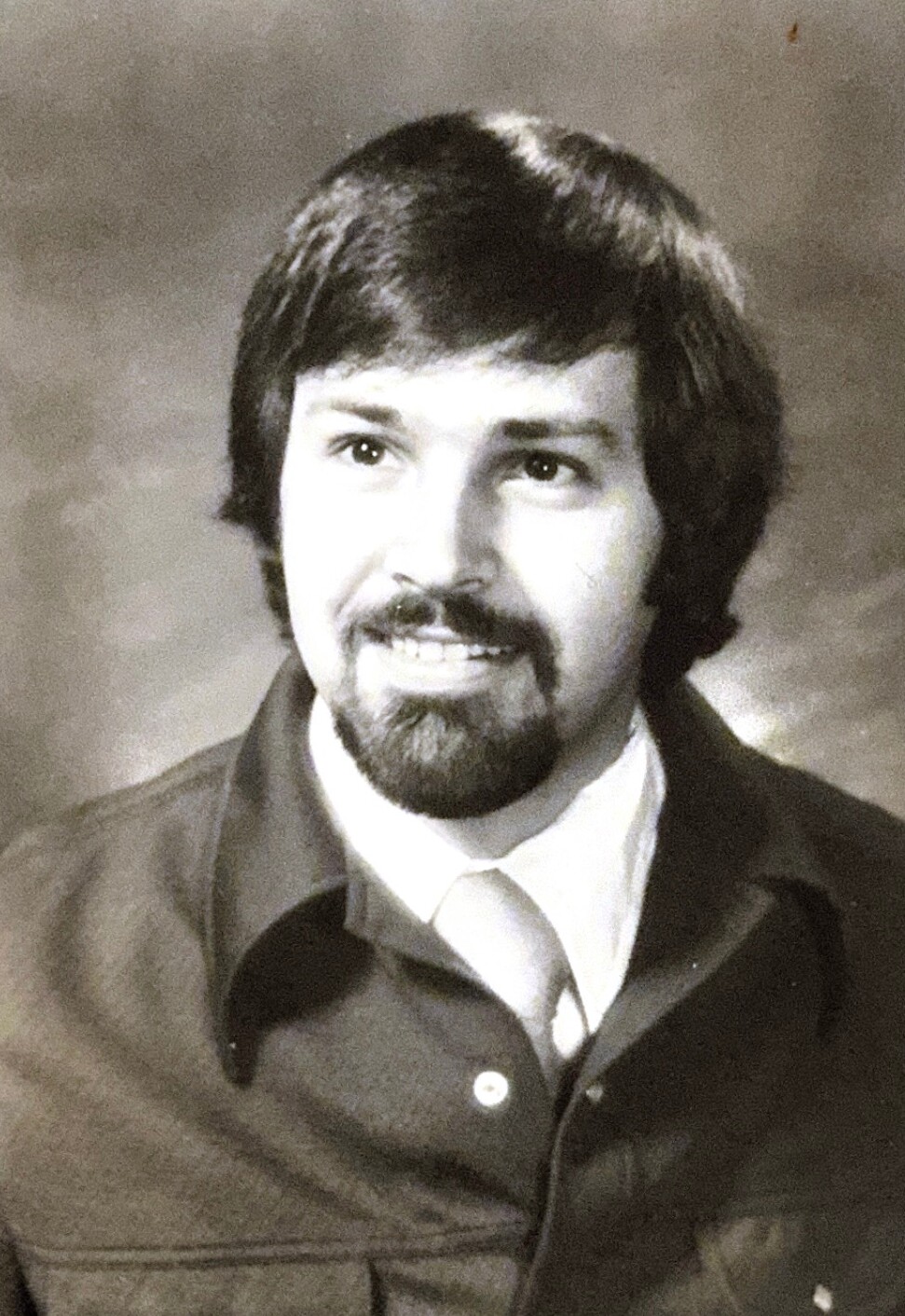 Black-and-white portrait of a man with dark hair and a beard, wearing a collared shirt and jacket, smiling softly.