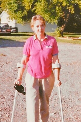 Woman outdoors, wearing a pink polo shirt, standing on gravel and using forearm crutches with braces on both arms.