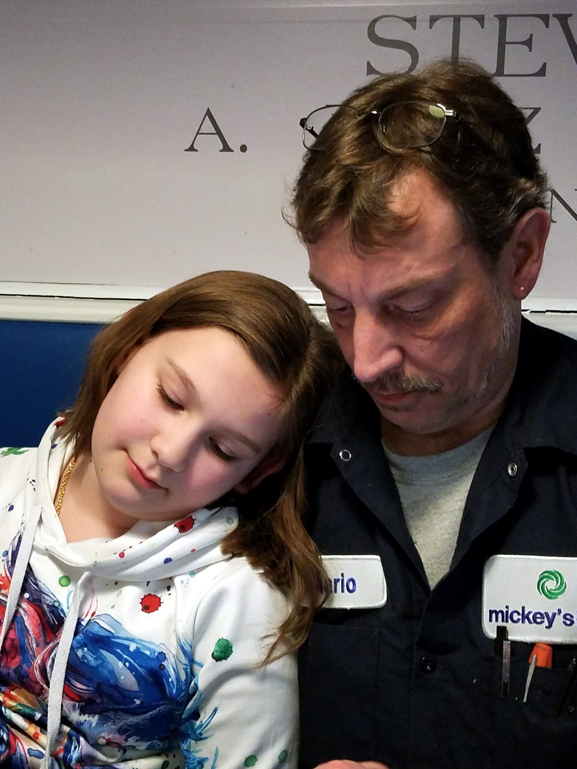Two people lean close together with eyes closed, smiling slightly; girl wears a colorful scarf and a white hoodie, man has glasses on his head and a name badge visible on his dark shirt.