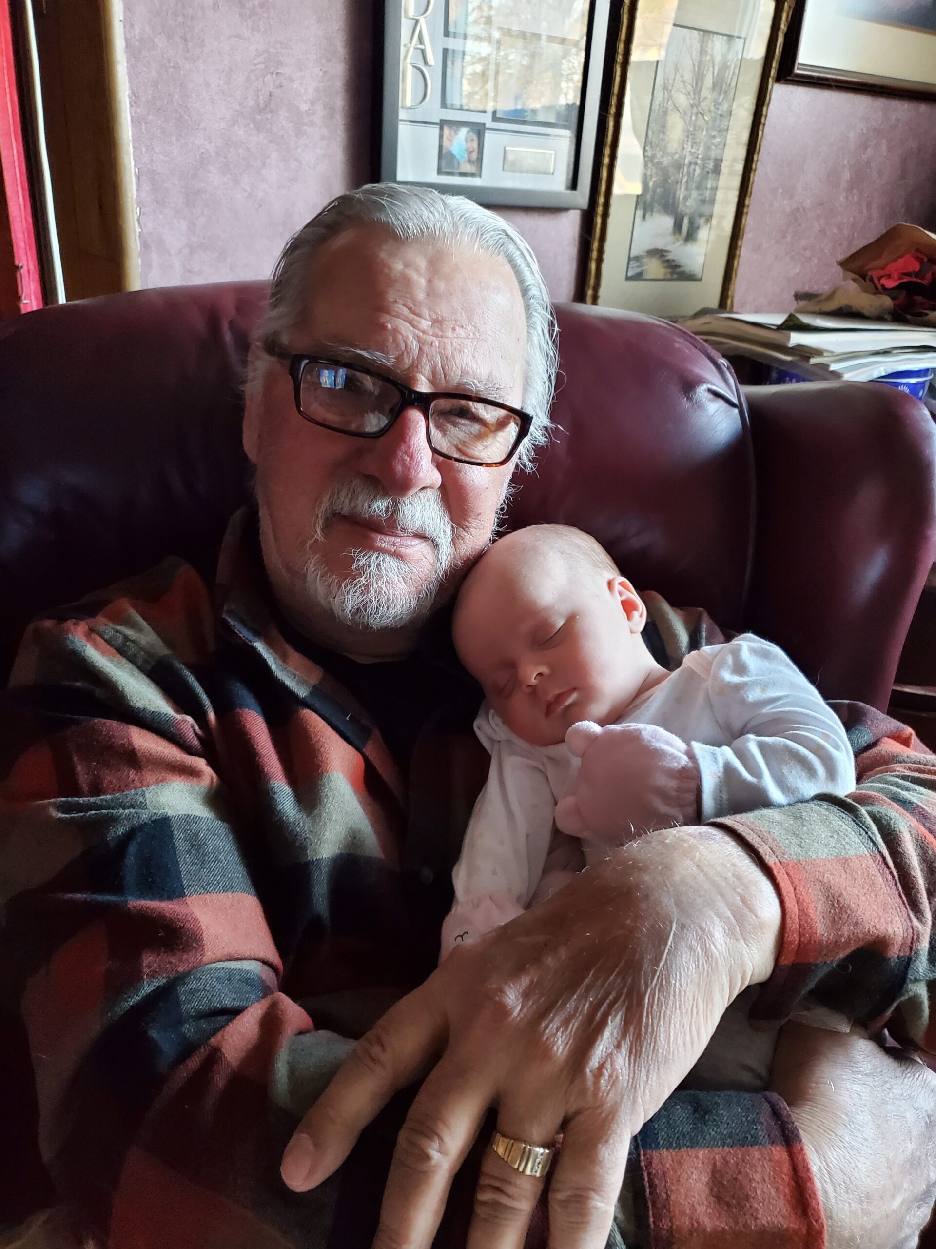Older man with glasses cradling a sleeping baby in his arms on a maroon leather chair inside a home.