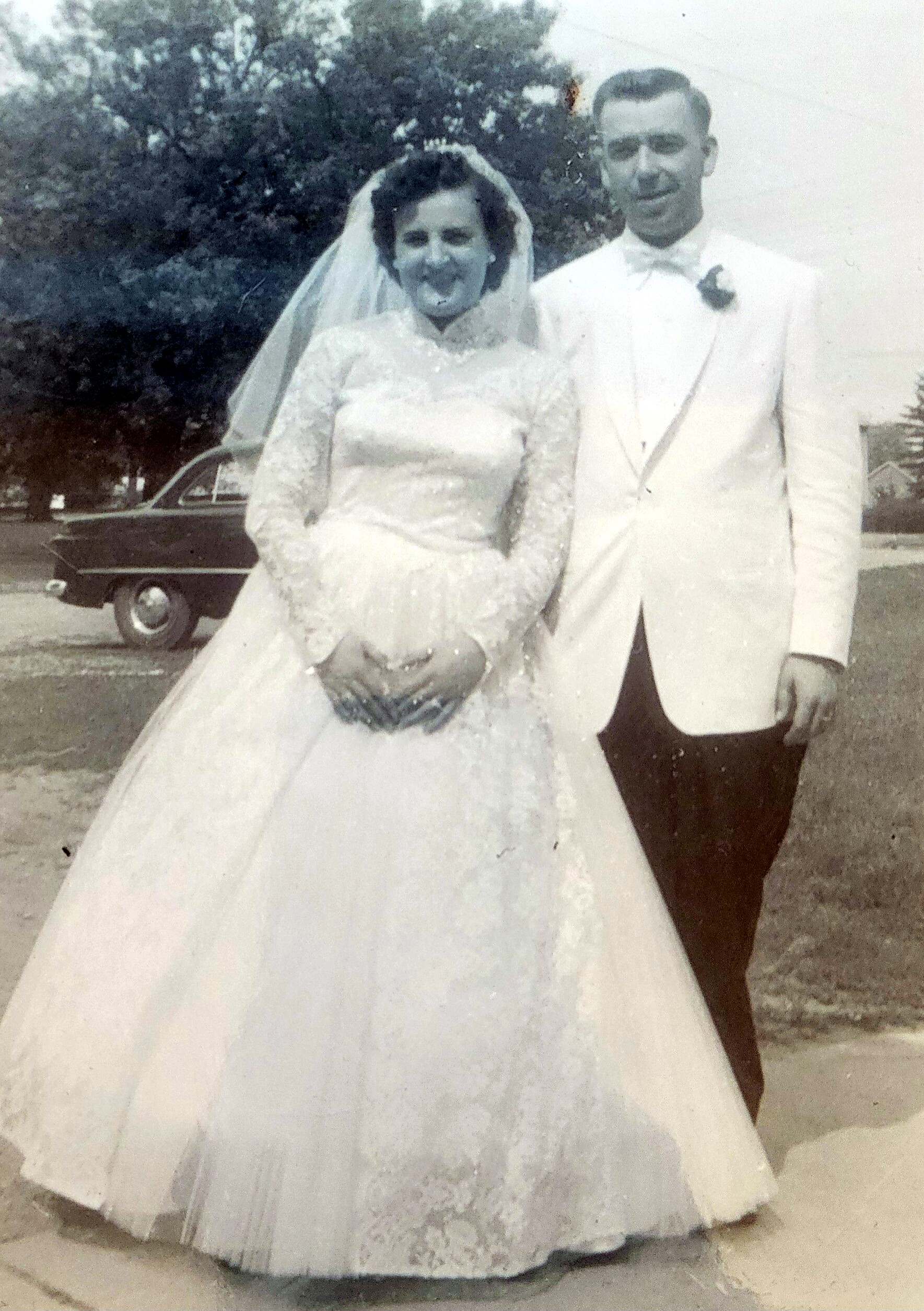 Bride in a lace wedding gown and veil smiles beside her groom in a light suit outdoors, a vintage car in the background.