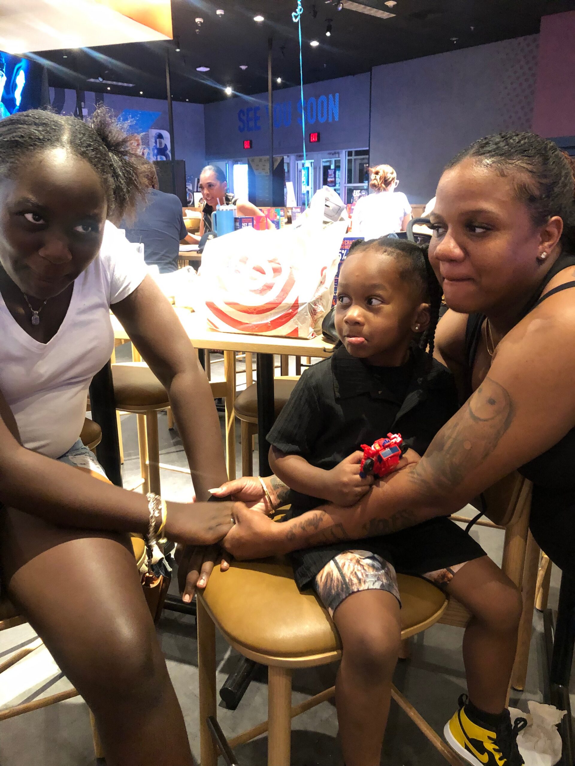 Three people sit at a counter in a casual restaurant; a woman in white leans in from left, a child sits with a toy car, and a tattooed woman on the right holds the child while smiling.