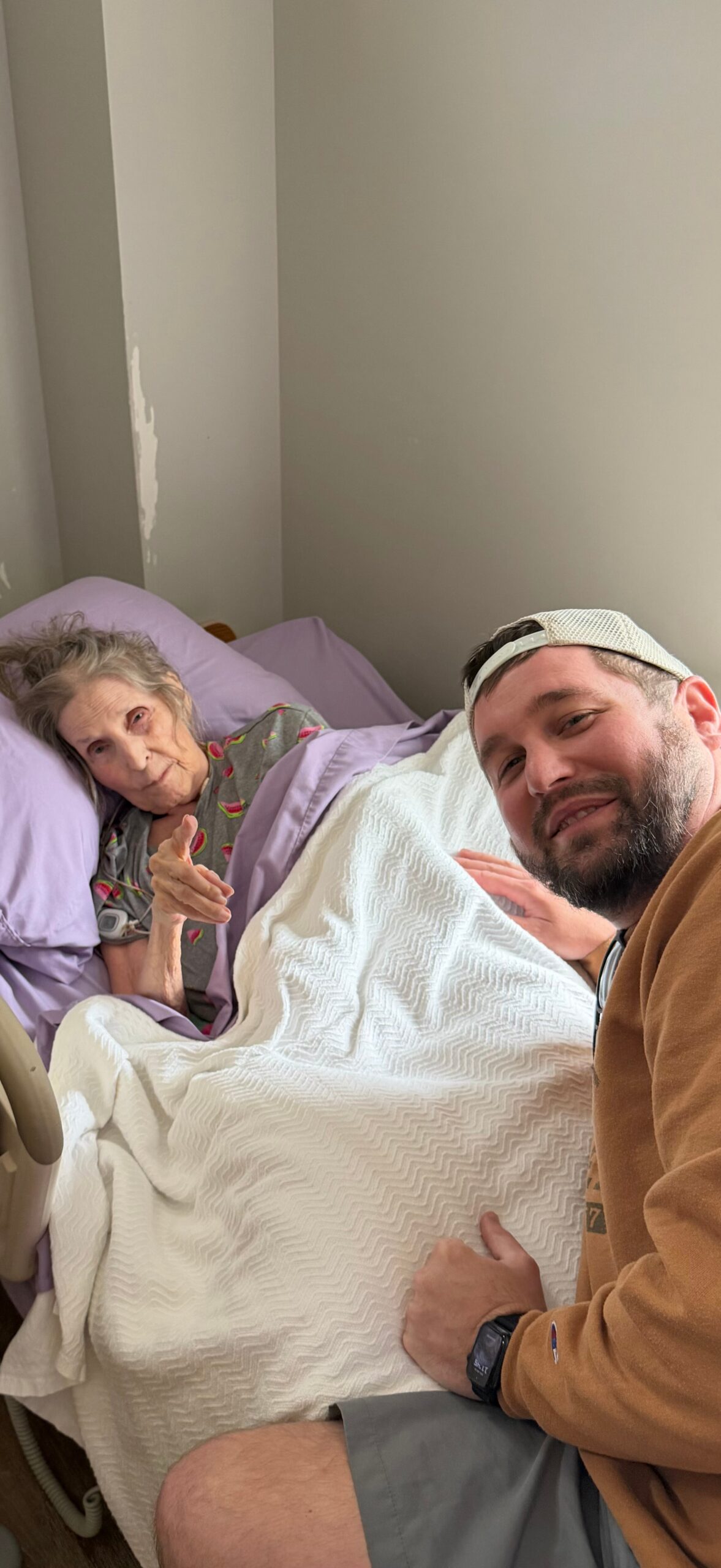 Smiling man in a brown jacket takes a selfie next to an elderly woman in a hospital bed, both under a white quilt.
