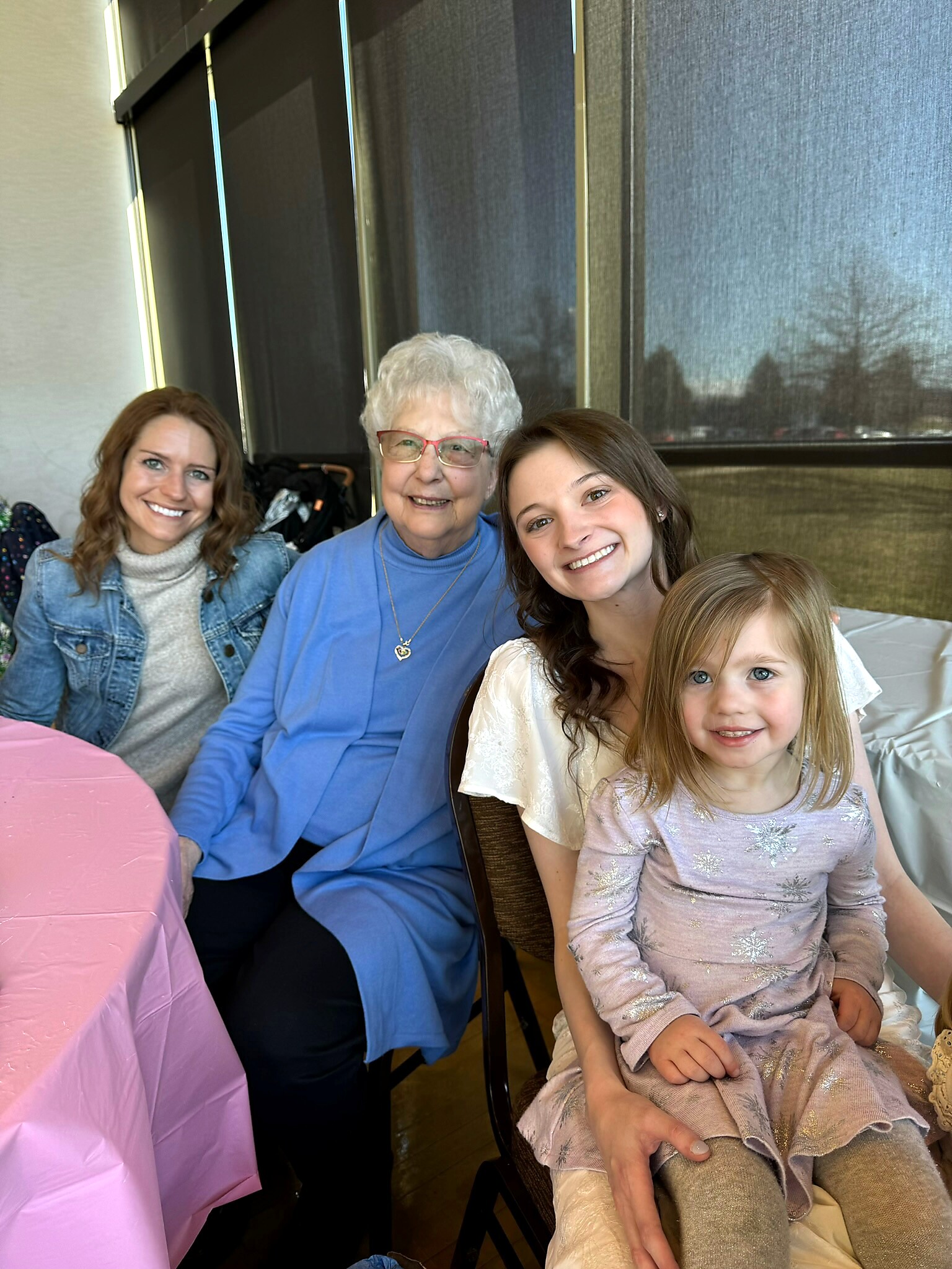 Smiling grandmother in a blue top with red glasses sits between two smiling younger women and a young girl in a sparkly dress at a table with a pink tablecloth.