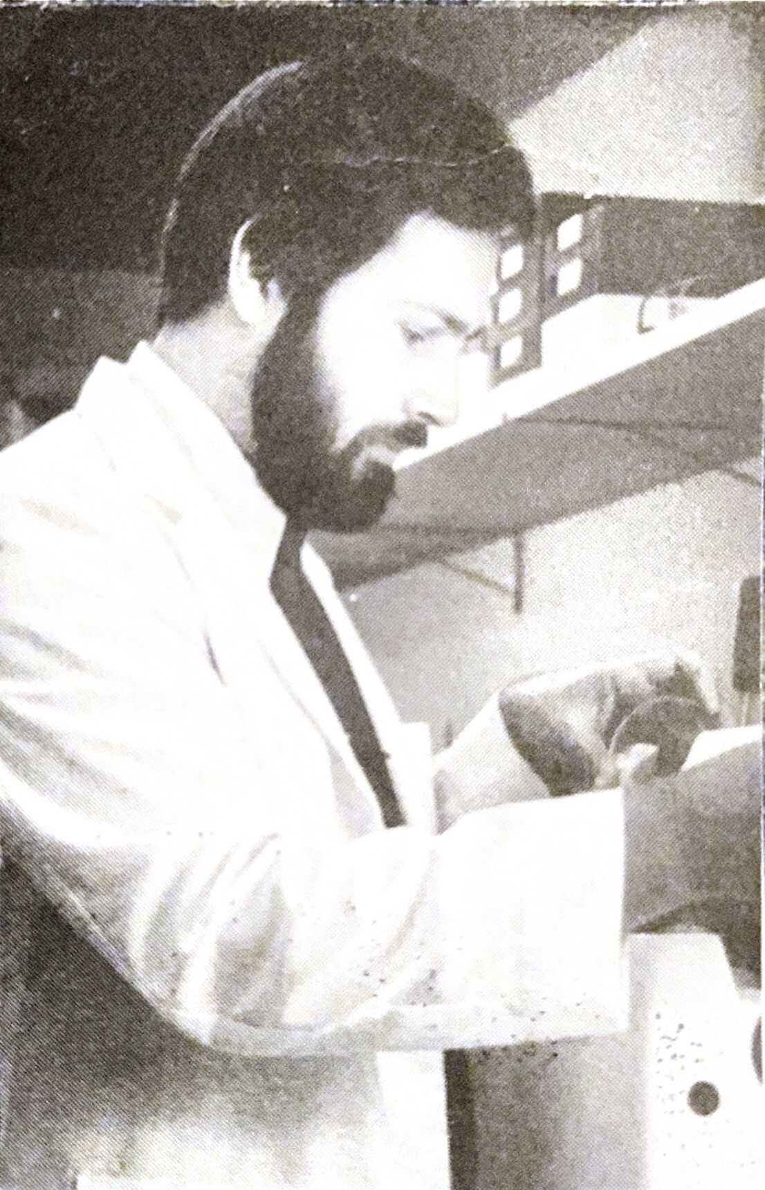 Man with dark hair and beard reads a document at a desk, shown in profile in a light-colored suit.