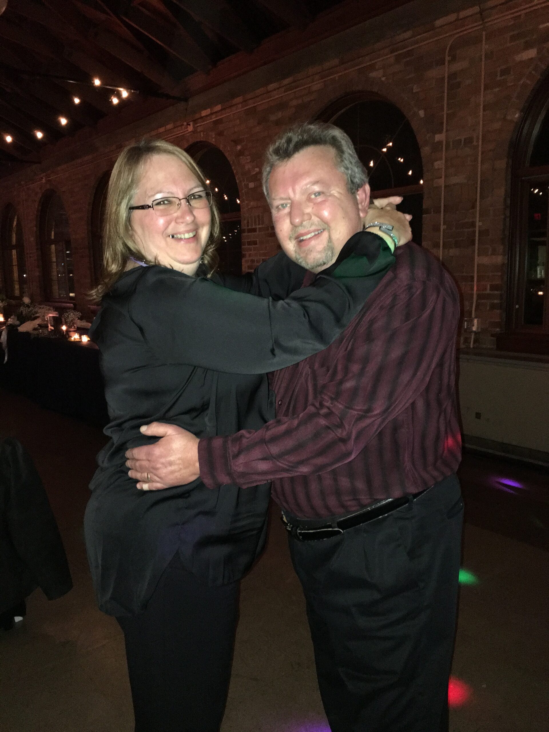 Couple dancing and embracing in a brick-walled, dimly lit venue with string lights, smiling at the camera.