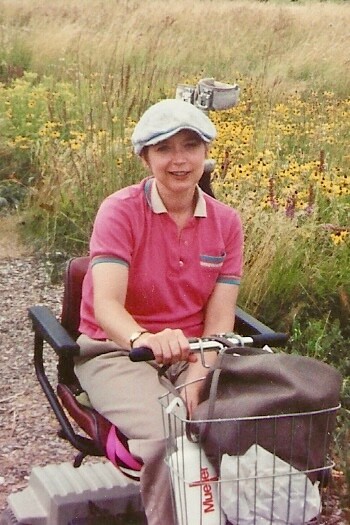 Woman in a pink polo shirt and light cap riding a bicycle with a front basket along a gravel path beside a wildflower field.