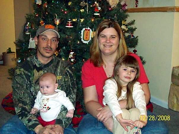 Family of four posing in front of a decorated Christmas tree: father in camo, mother in red, young daughter in white, baby son on his lap.