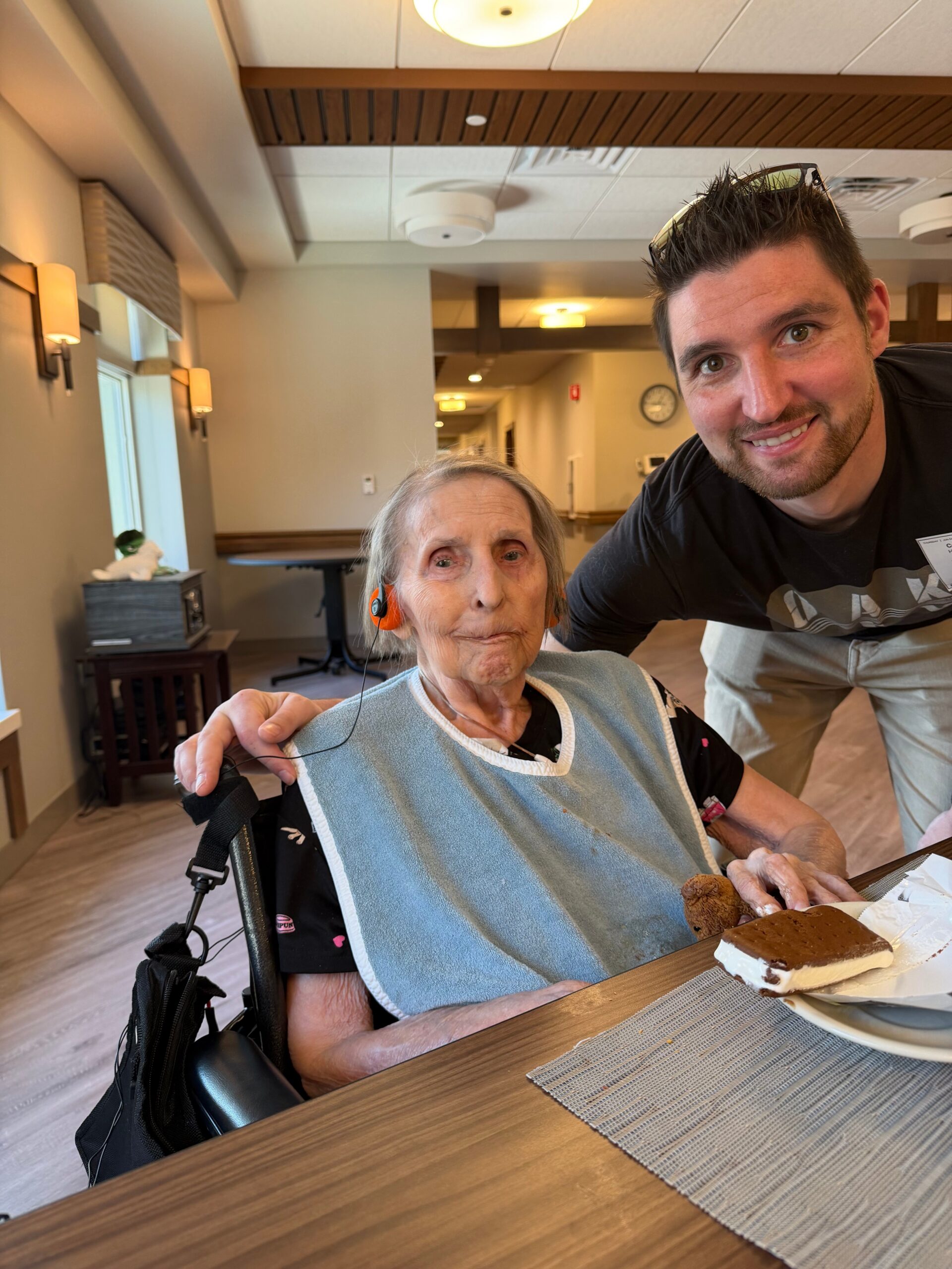 Smiling elderly woman in a wheelchair wearing a blue bib sits at a dining table with a slice of chocolate cake on her plate as a man leans in from the right for a selfie in a care-home dining area.