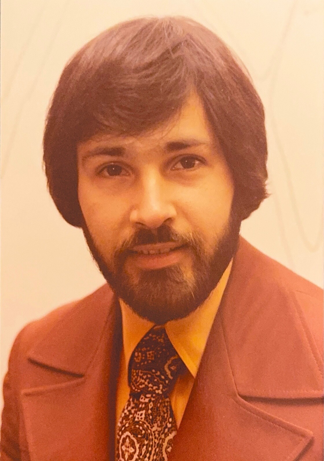 Portrait of a man with dark hair and beard wearing a brown suit, yellow shirt, and paisley tie, looking at the camera.