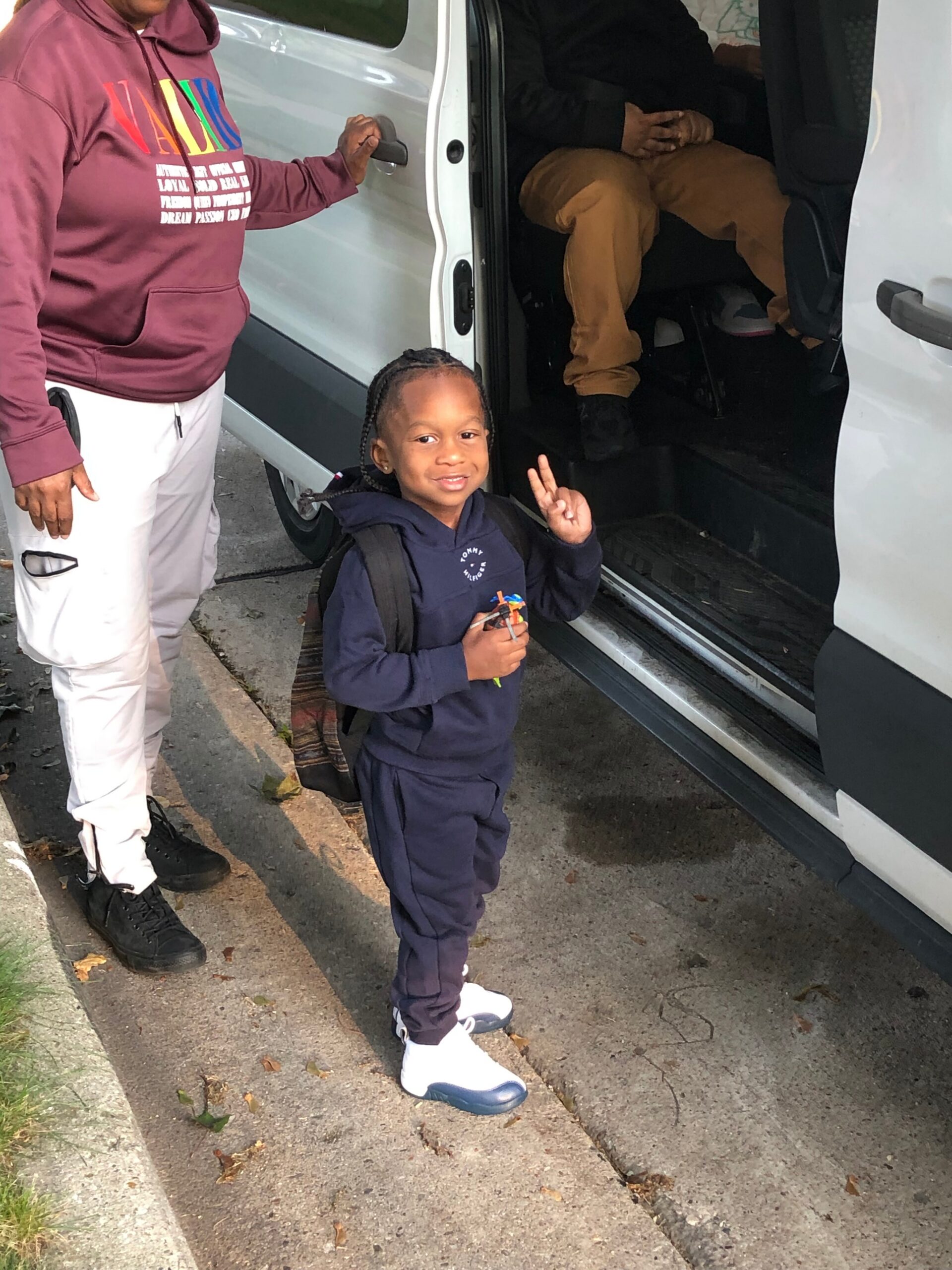 Young child in navy tracksuit with a backpack stands on the sidewalk beside an open white van, flashing a peace sign, while an adult in a maroon hoodie stands nearby.