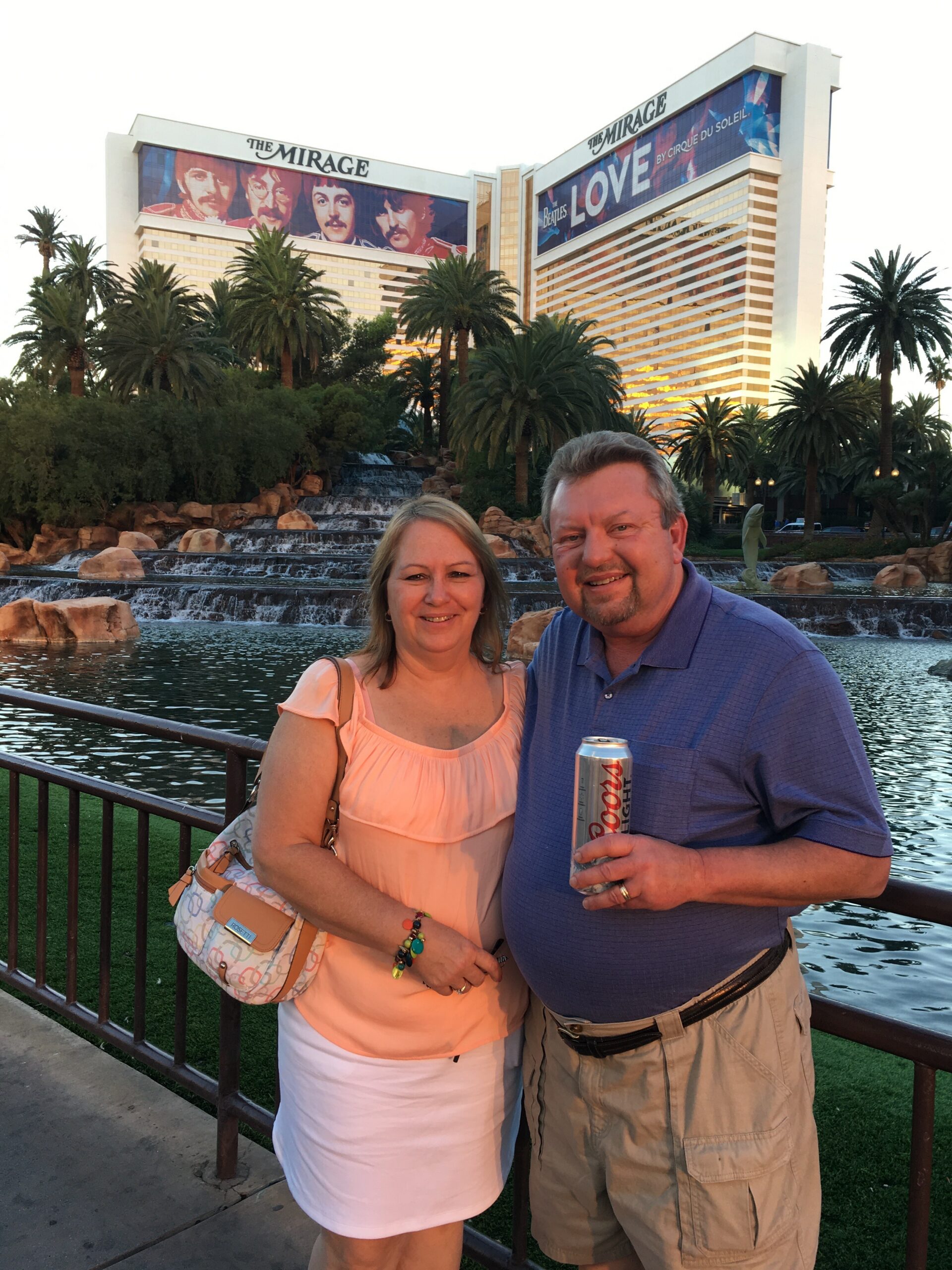 Smiling couple standing by a railing in front of The Mirage in Las Vegas, with a tiered waterfall behind them and palm trees nearby.