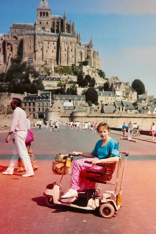 Person on a mobility scooter rides along a riverside promenade, with a medieval fortress perched on a hill in the background.