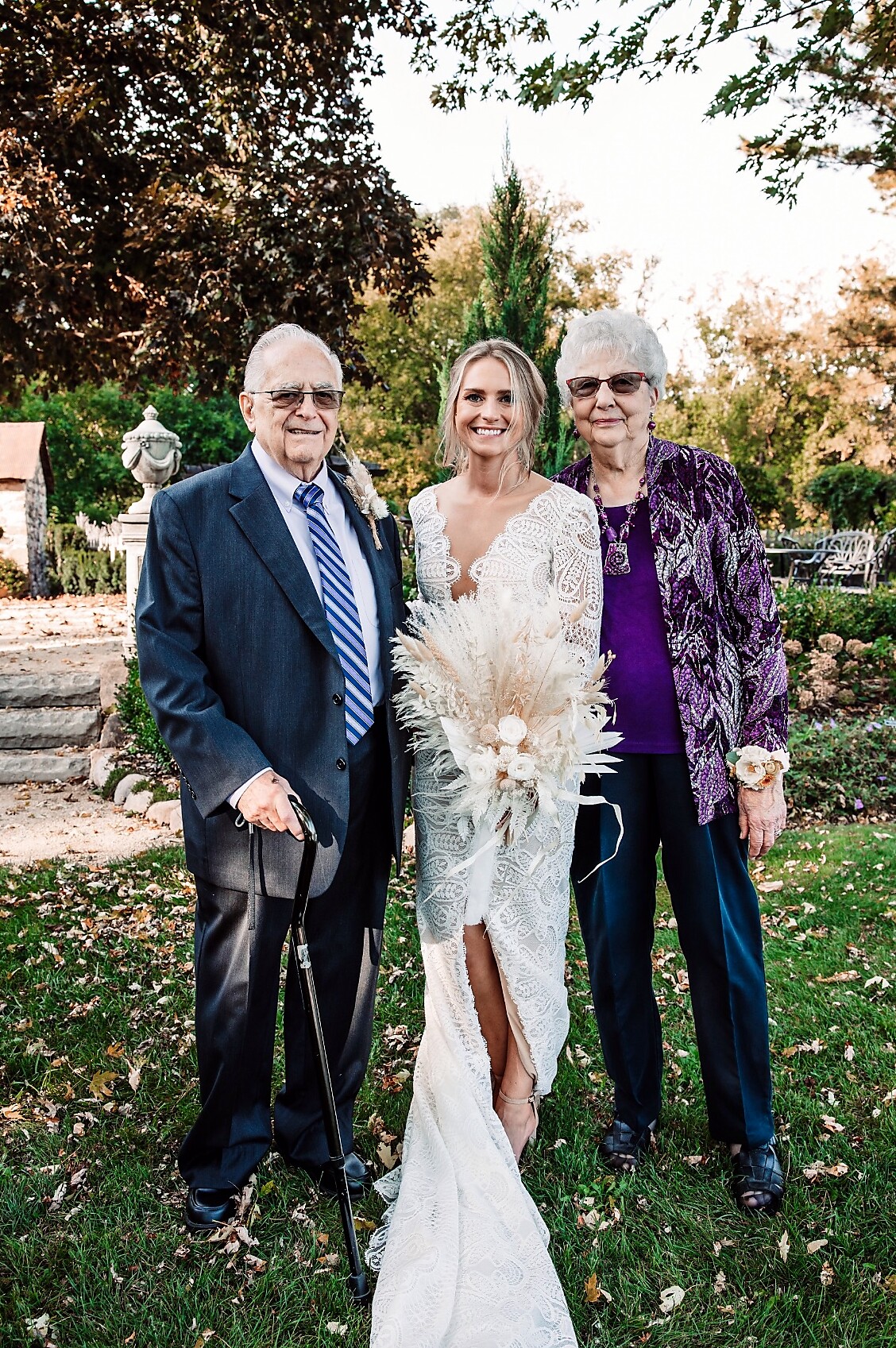 Bride in a lace wedding dress holding a pale bouquet, flanked by an older man in a suit and an older woman in a purple jacket outdoors at a garden wedding.