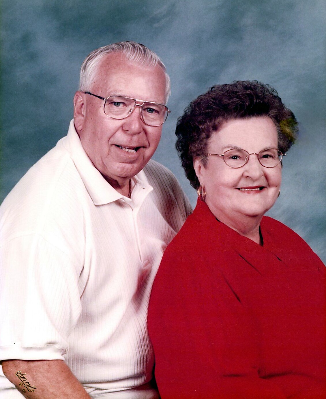Smiling elderly couple posing for a portrait; man in a white polo and woman in a red blazer wearing glasses.