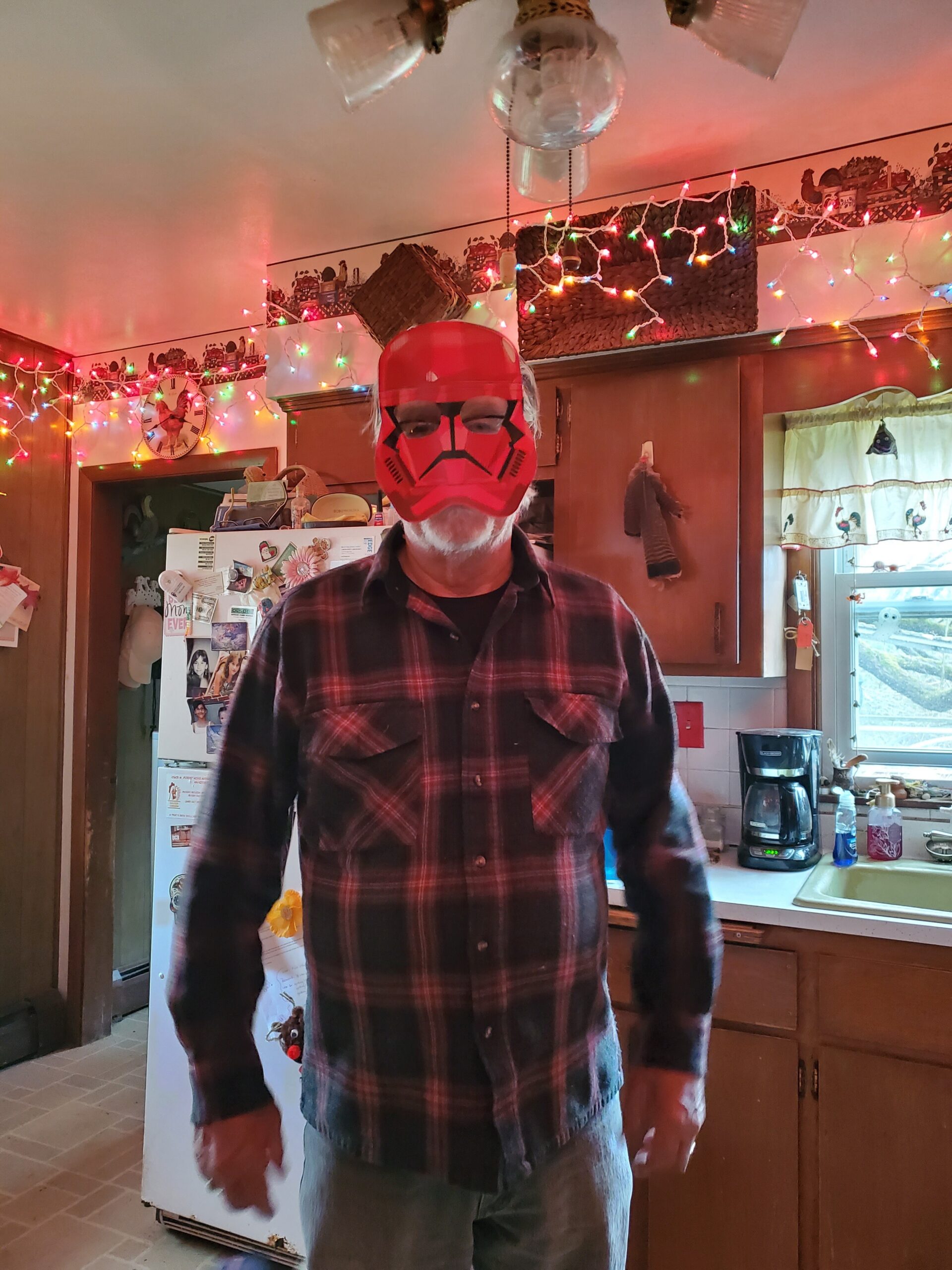 Man wearing a red helmet-style mask indoors in a kitchen, standing in front of a fridge and colorful string lights overhead.