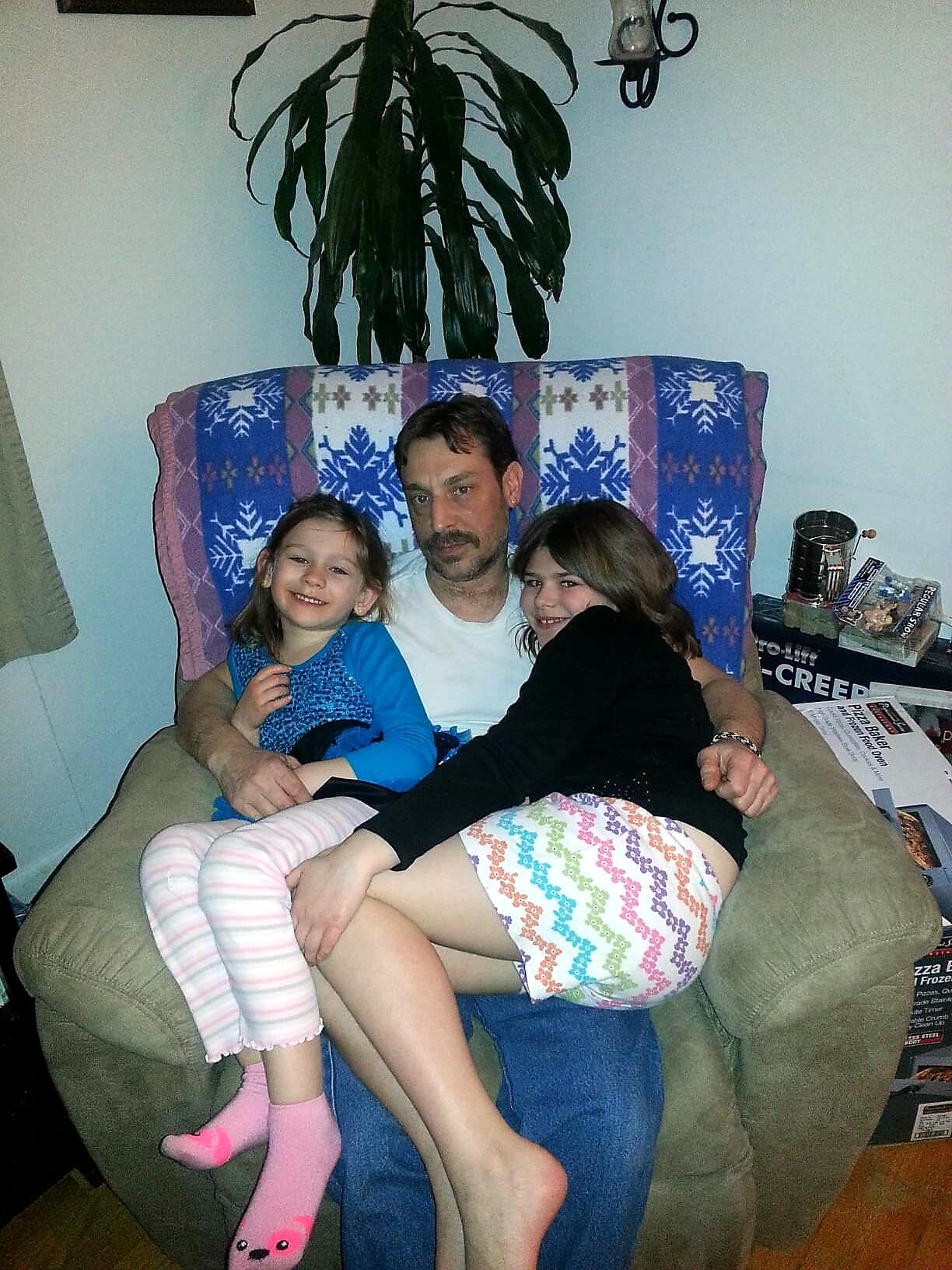 Father sits on a couch with two young girls cuddled beside him, in a living room with a large plant behind them and a blue snowflake blanket on the backrest