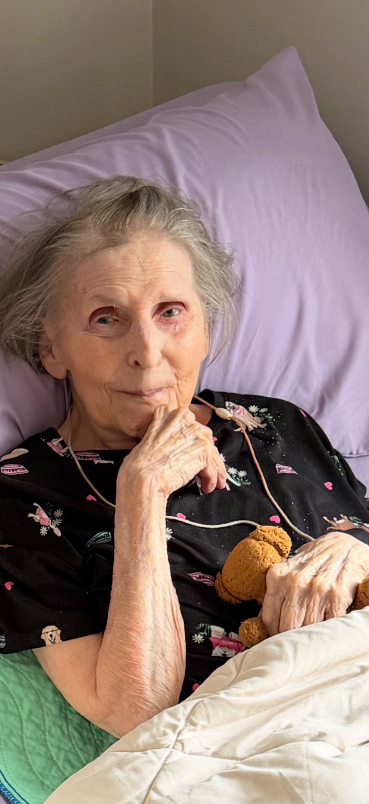 Older woman resting in bed on a purple pillow, wearing a black printed gown and holding a brown stuffed bear, with a gentle smile.
