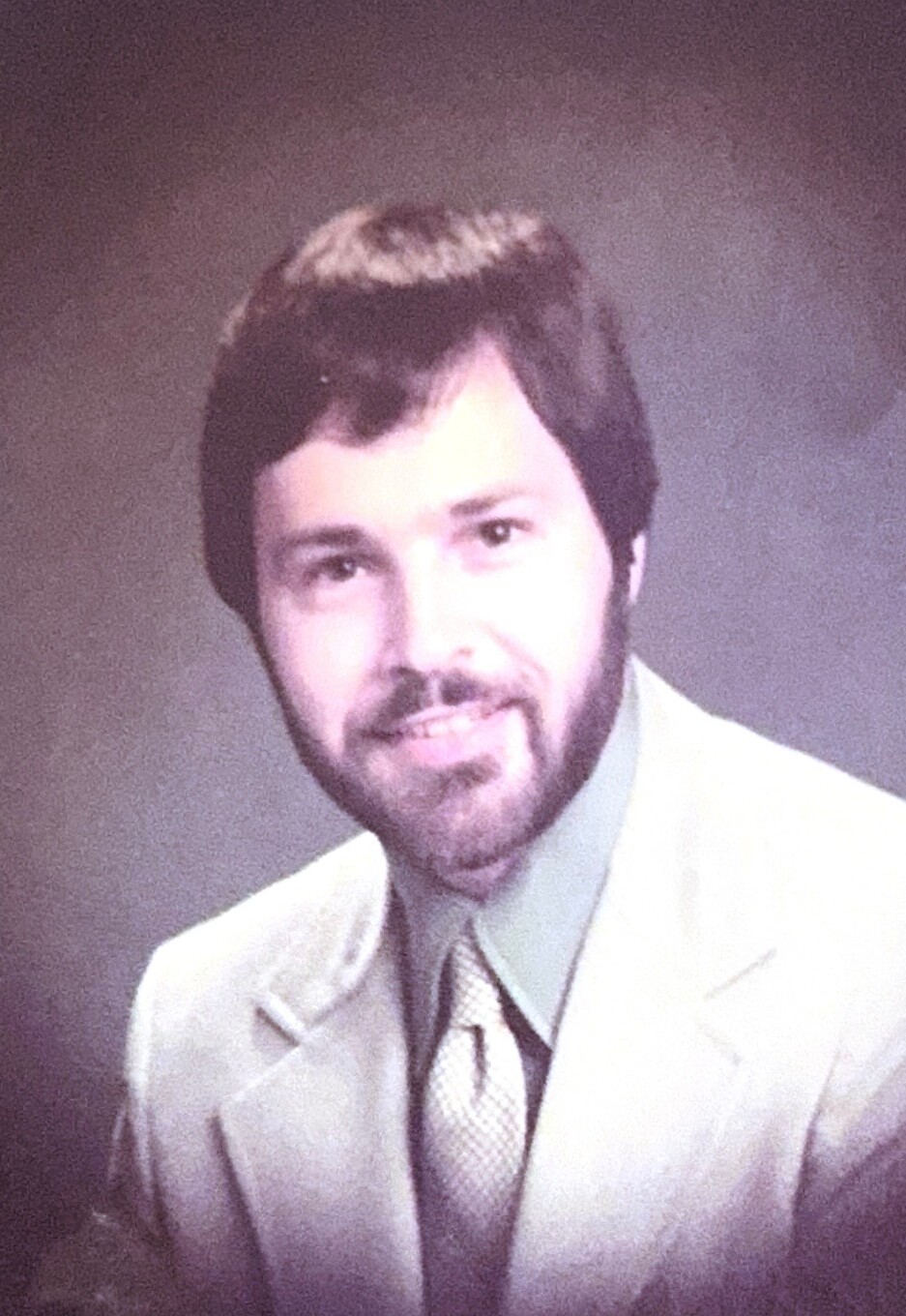 Portrait of a man with dark hair and beard, wearing a light suit and tie, smiling softly in a studio portrait.