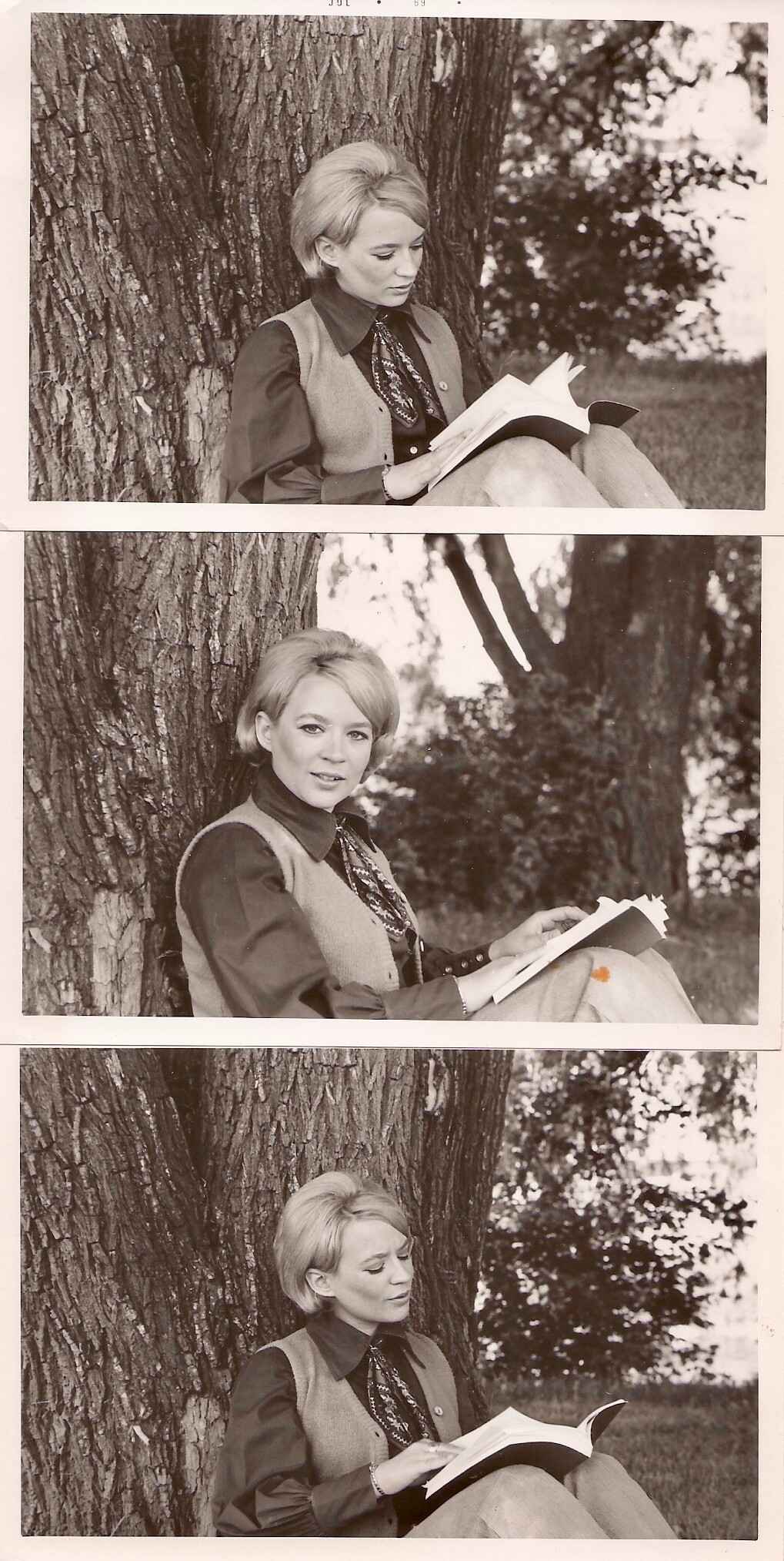 Woman with short blonde hair sits against a tree reading a notebook outdoors in a park setting.
