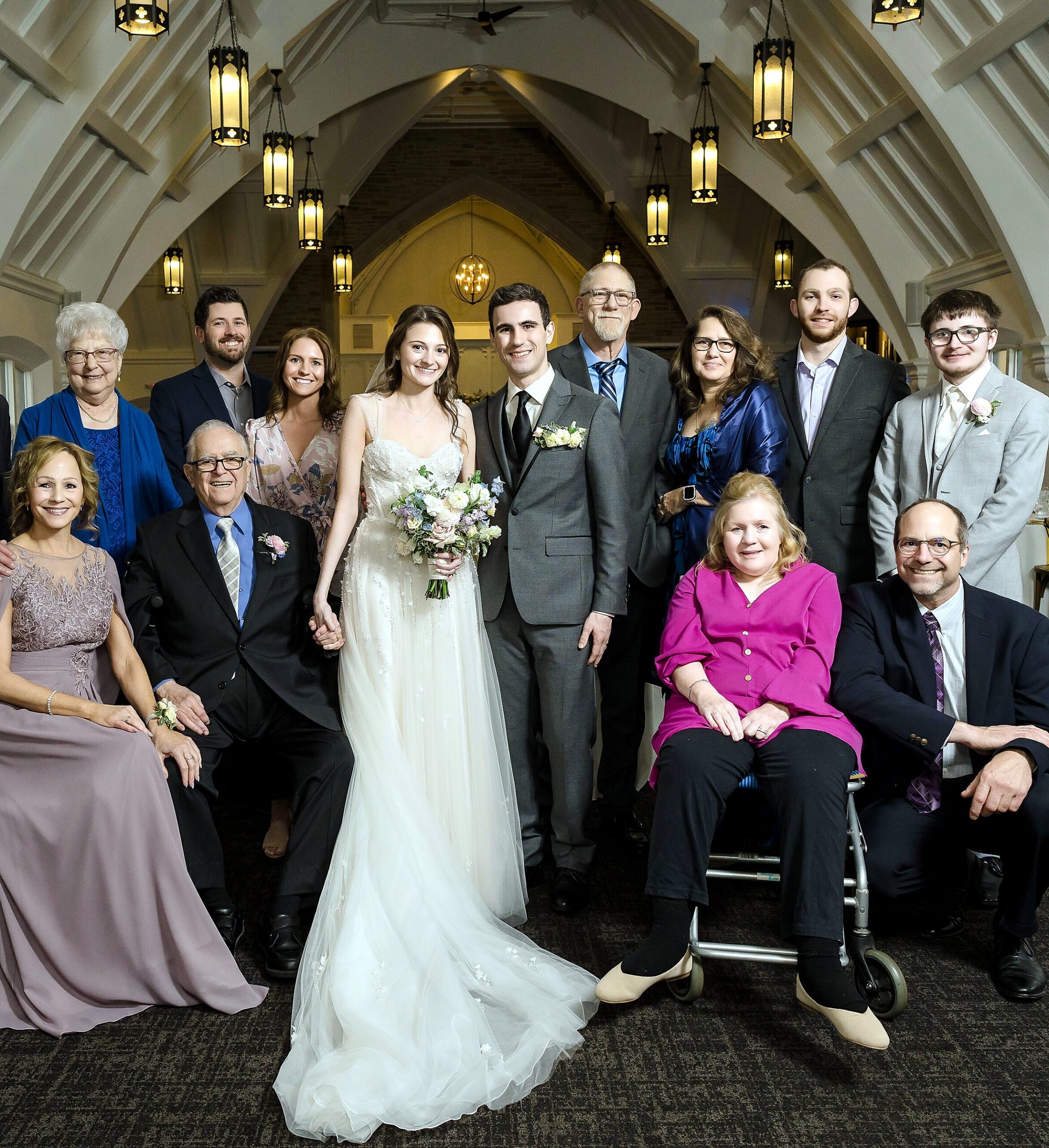 Bride in a white wedding gown and groom in a gray suit pose with family in a church foyer under hanging lanterns to the left and right.