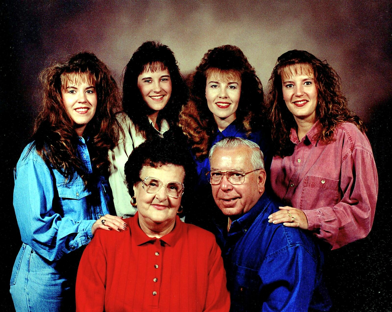 Group portrait of six family members: an older couple seated in front with four smiling daughters standing behind them in colorful shirts.