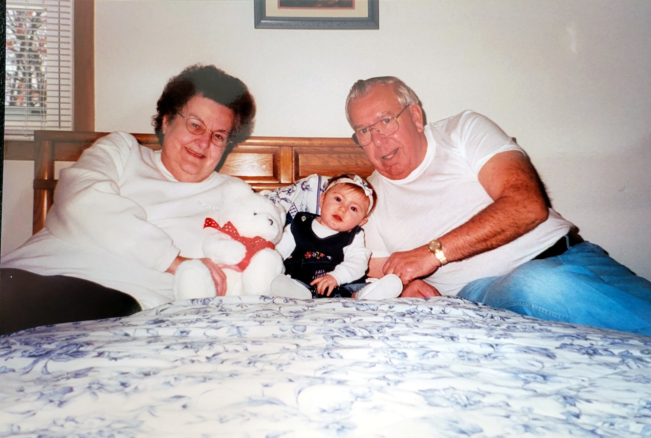 Grandparents and baby sitting together on a bed, smiling at the camera in a cozy family moment