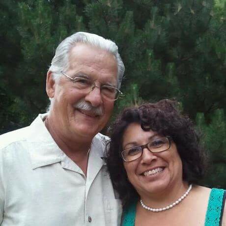 Smiling older couple posing outdoors with green trees in the background, close together in portrait.