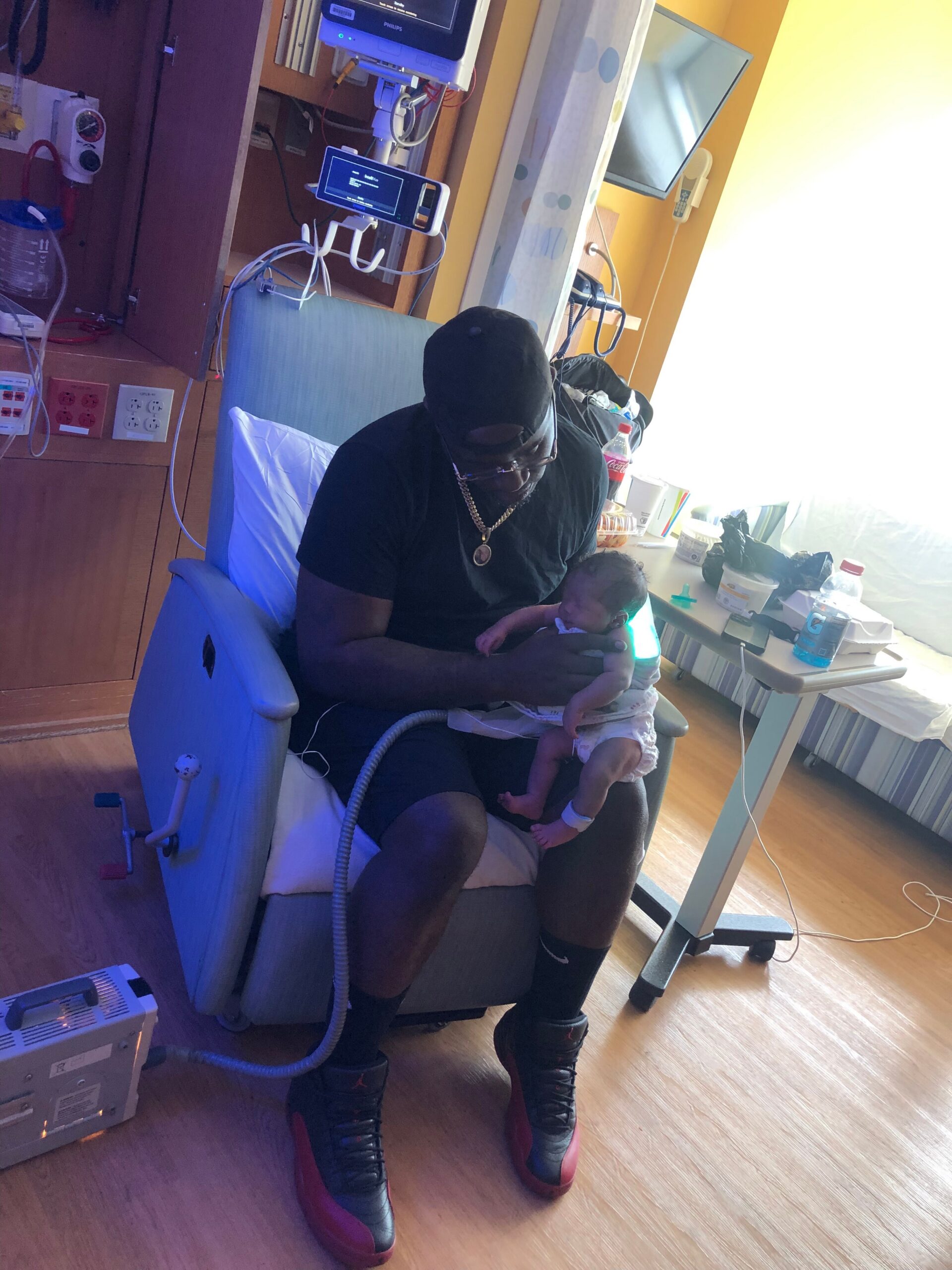 Man in a black cap and gold chain sits in a chair, cradling a newborn baby beside a hospital bed with medical equipment nearby.