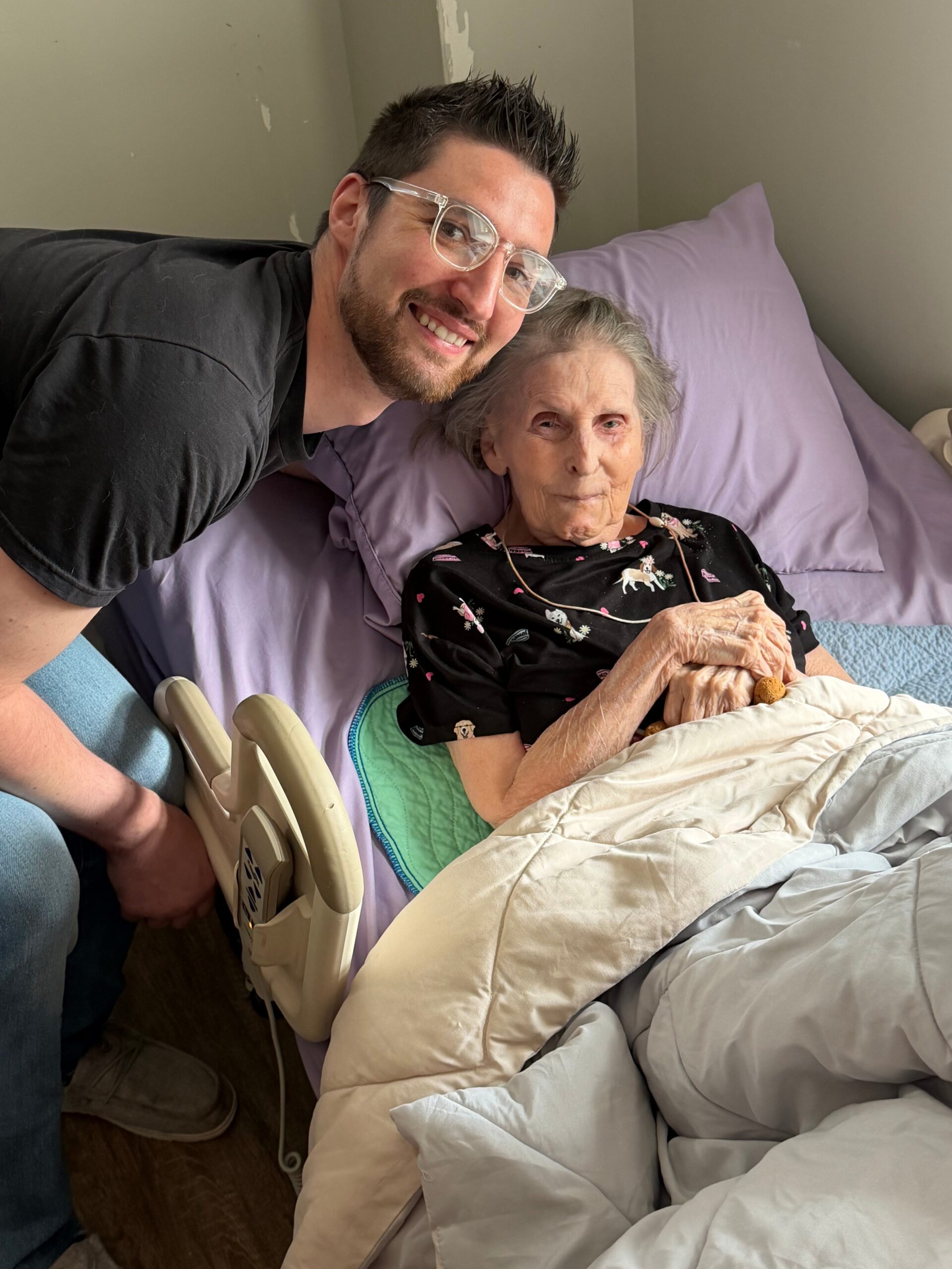 Smiling man with clear glasses leaning over an elderly woman in bed, holding her hand in a caregiving moment.