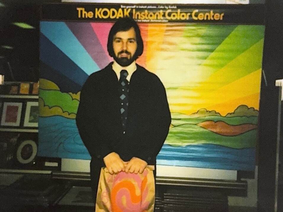 Man in a dark suit and tie stands in front of a rainbow mural with a Kodak Instant Color Center sign, holding a pink and orange patterned bag.